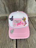A pink and white trucker hat with western-themed embroidery, rhinestone trim, and decorative patches, displayed on a rustic wooden surface.