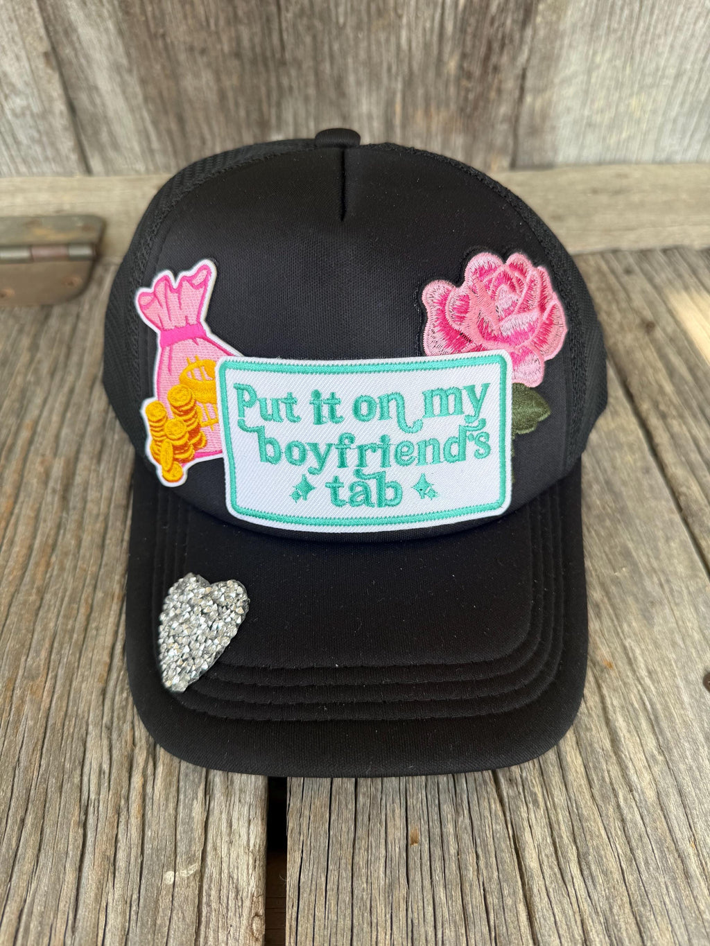 A black trucker-style cap with decorative embroidered patches, including a pink rose, a pink and yellow bunny, a silver rhinestone heart, and a central patch that reads 'Put it on my boyfriend's tab' in teal lettering on a white background. The hat is photographed on a rustic wooden surface.