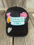 A black trucker-style cap with decorative embroidered patches, including a pink rose, a pink and yellow bunny, a silver rhinestone heart, and a central patch that reads 'Put it on my boyfriend's tab' in teal lettering on a white background. The hat is photographed on a rustic wooden surface.
