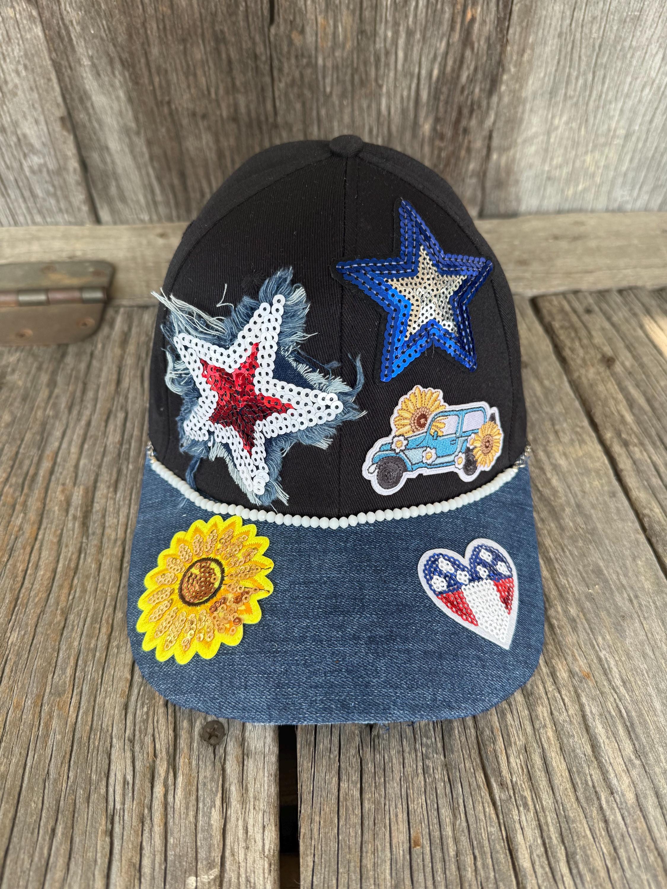 A black baseball cap with a blue denim brim, decorated with multiple western-style embellishments including rhinestone stars, a sunflower patch, a car patch, and a heart patch, displayed on a rustic wooden surface.