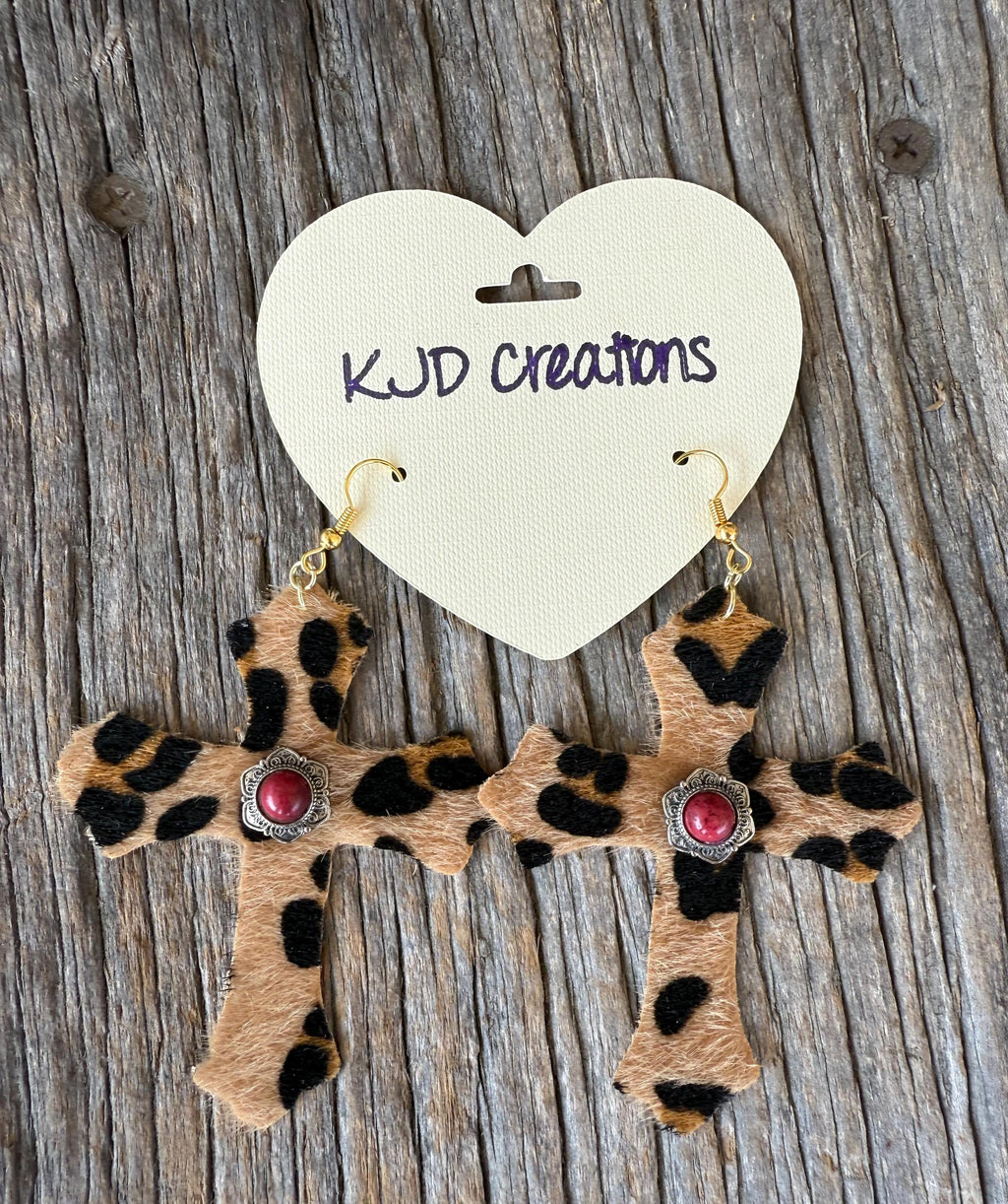 A pair of leopard-print cross dangle earrings with red gemstone centers and silver-toned settings, hanging from gold hooks on a cream heart-shaped card that reads 'KJD Creations,' placed on a weathered wood surface.