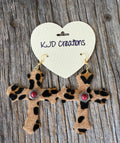 A pair of leopard-print cross dangle earrings with red gemstone centers and silver-toned settings, hanging from gold hooks on a cream heart-shaped card that reads 'KJD Creations,' placed on a weathered wood surface.