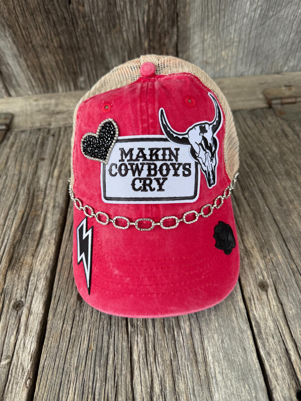 A red and beige trucker-style baseball cap with western-themed patches, including a cow skull, a black heart, a lightning bolt, and a chain detail across the brim, 