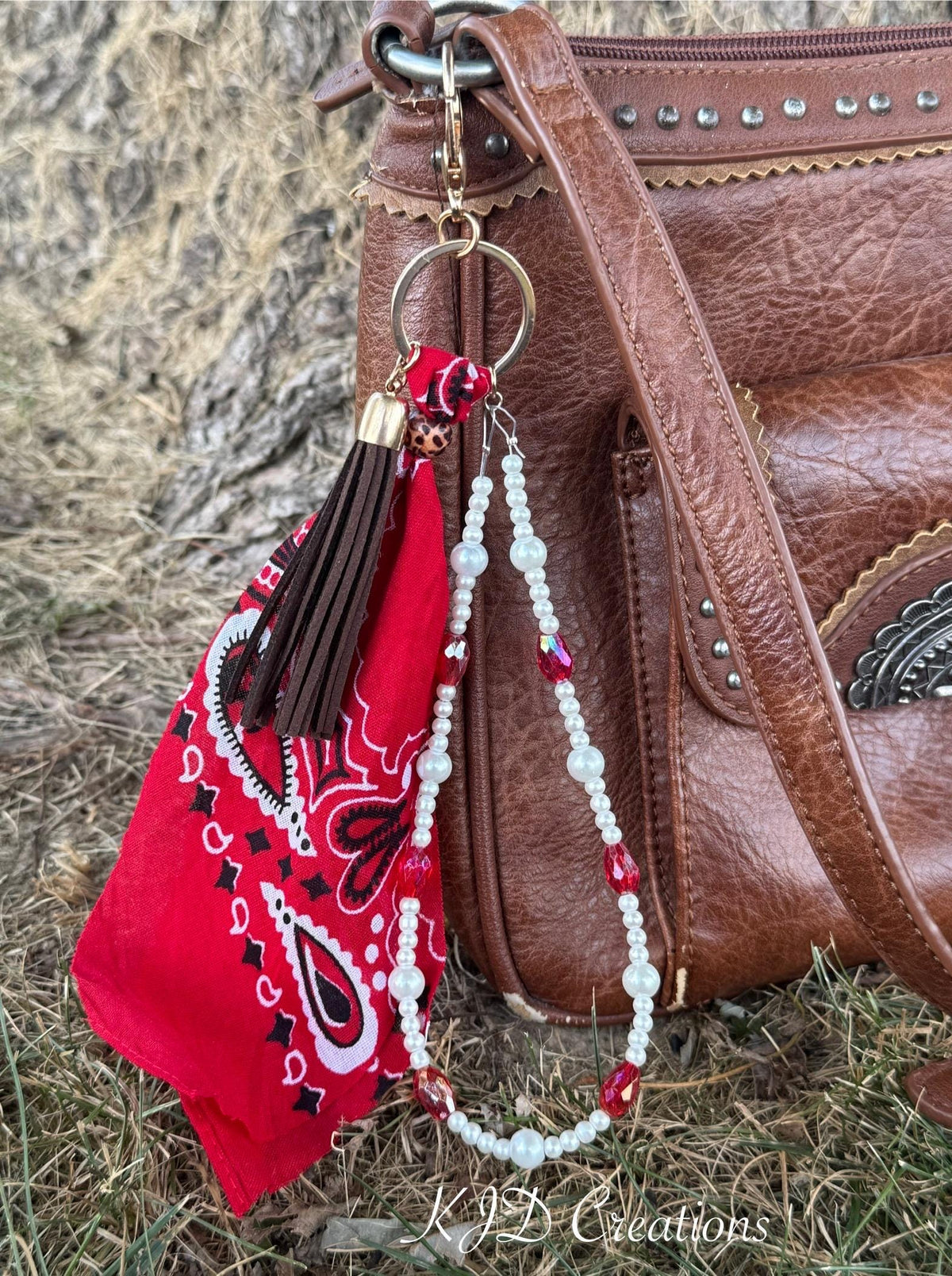Close-up of red paisley bandana bag charm on brown distressed studded Western leather crossbody bag showing gold lobster clasp and ring, red bandana, dark brown leather fringe tassel with leopard print bead cap, and double strand of white faux pearl beads with red AB crystal bead accents