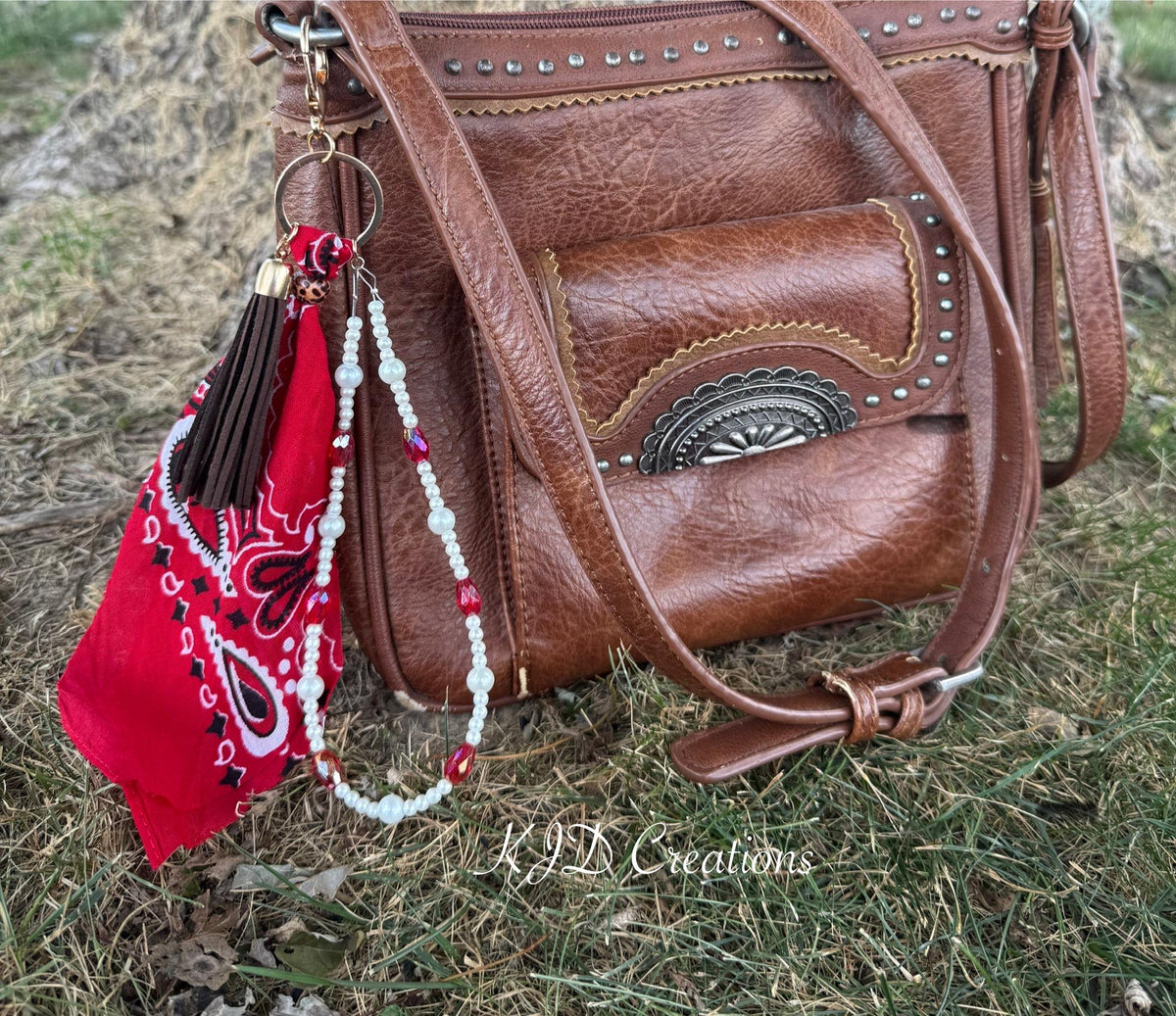 Side view of brown distressed studded Western leather crossbody bag on grass with red paisley bandana bag charm clipped to hardware featuring dark brown leather fringe tassel with leopard print bead cap and double strand of white faux pearl beads with red AB crystal bead accents