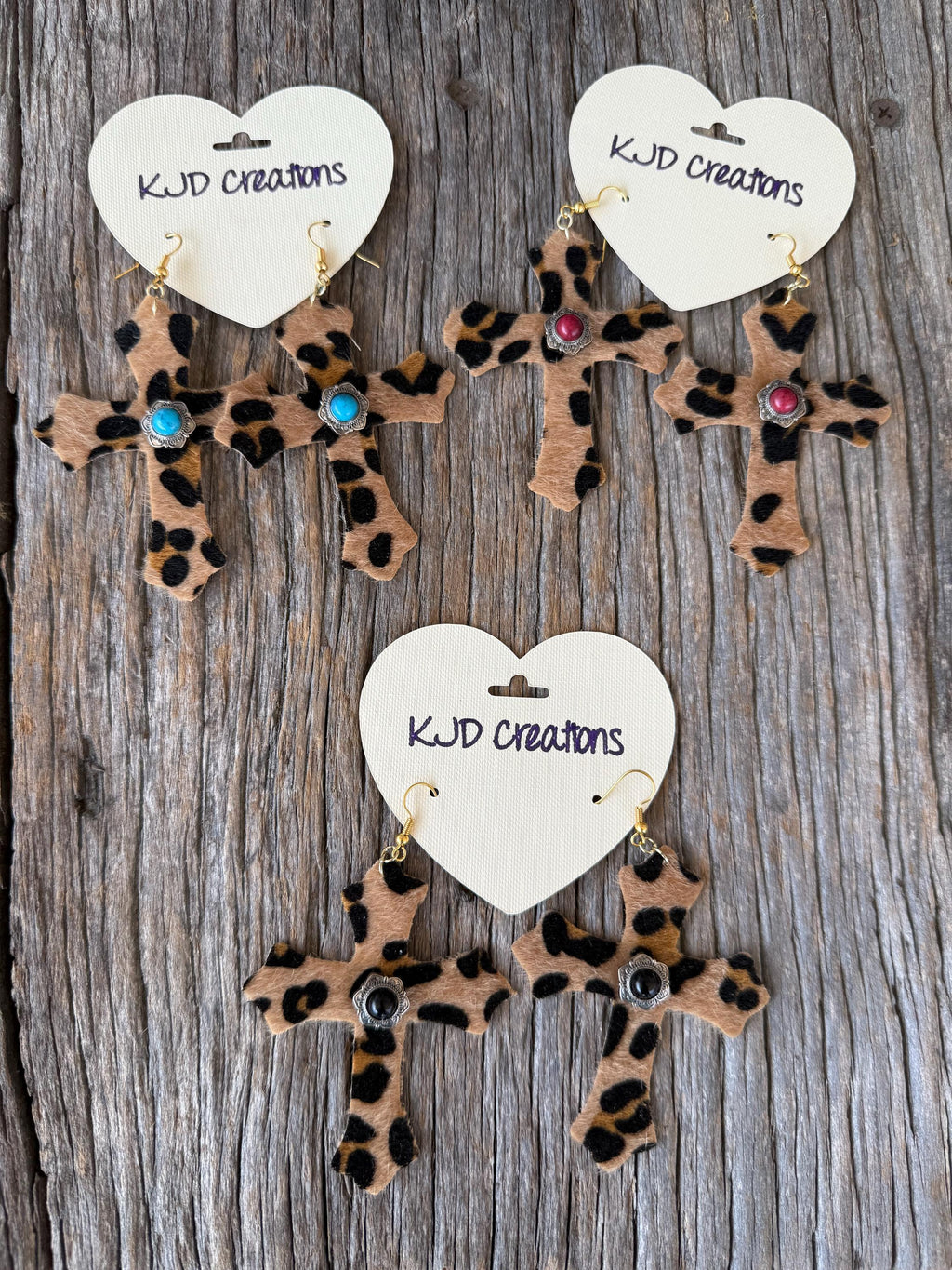 Three leopard-print cross dangle earrings with gold-tone hooks and colorful rhinestones, displayed on white heart-shaped cards labeled 'KJD creations' on a weathered wood background.
