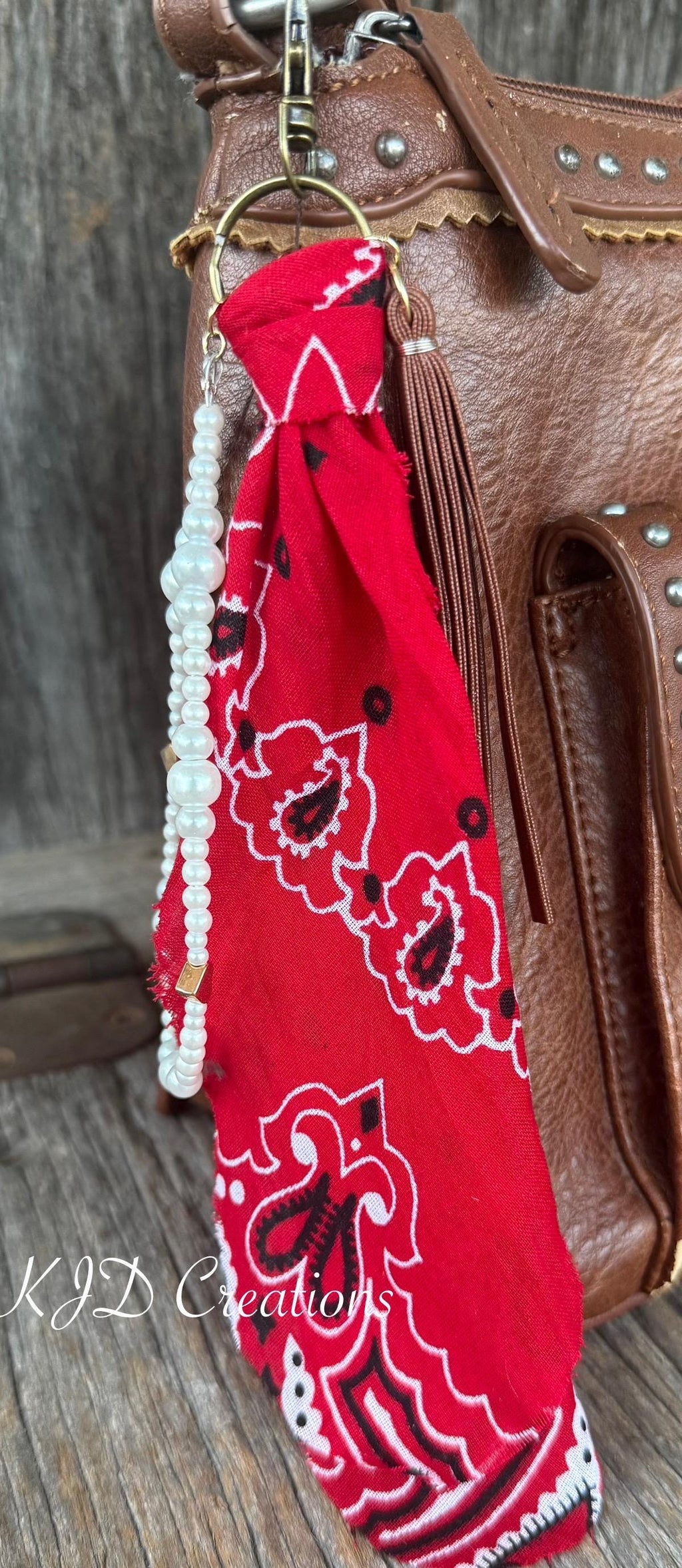 Close-up of red paisley bandana bag charm on brown studded Western leather crossbody bag against barn wood showing gold lobster clasp and ring, red bandana, white faux pearl bead strand with gold heart-shaped bead, and brown leather fringe tassel