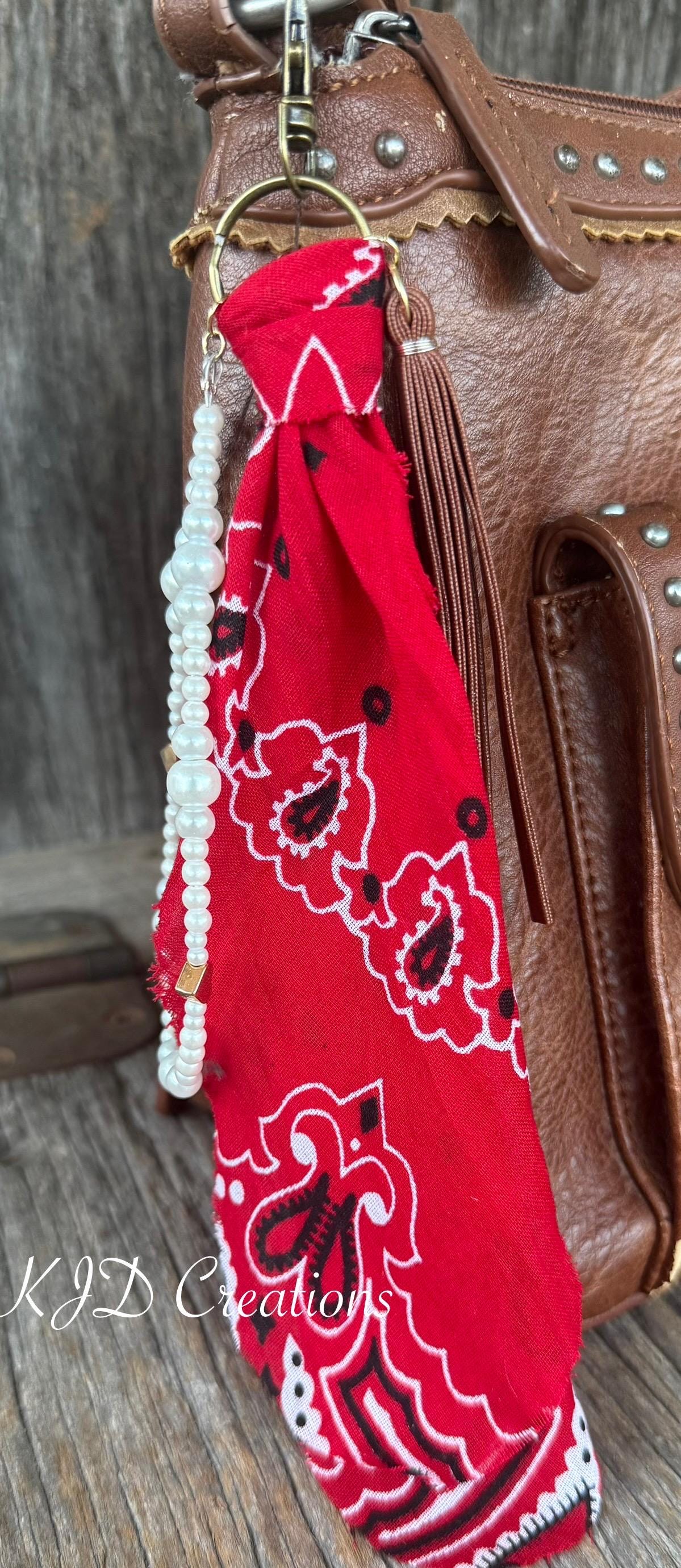 Close-up of red paisley bandana bag charm on brown studded Western leather crossbody bag against barn wood showing gold lobster clasp and ring, red bandana, white faux pearl bead strand with gold heart-shaped bead, and brown leather fringe tassel