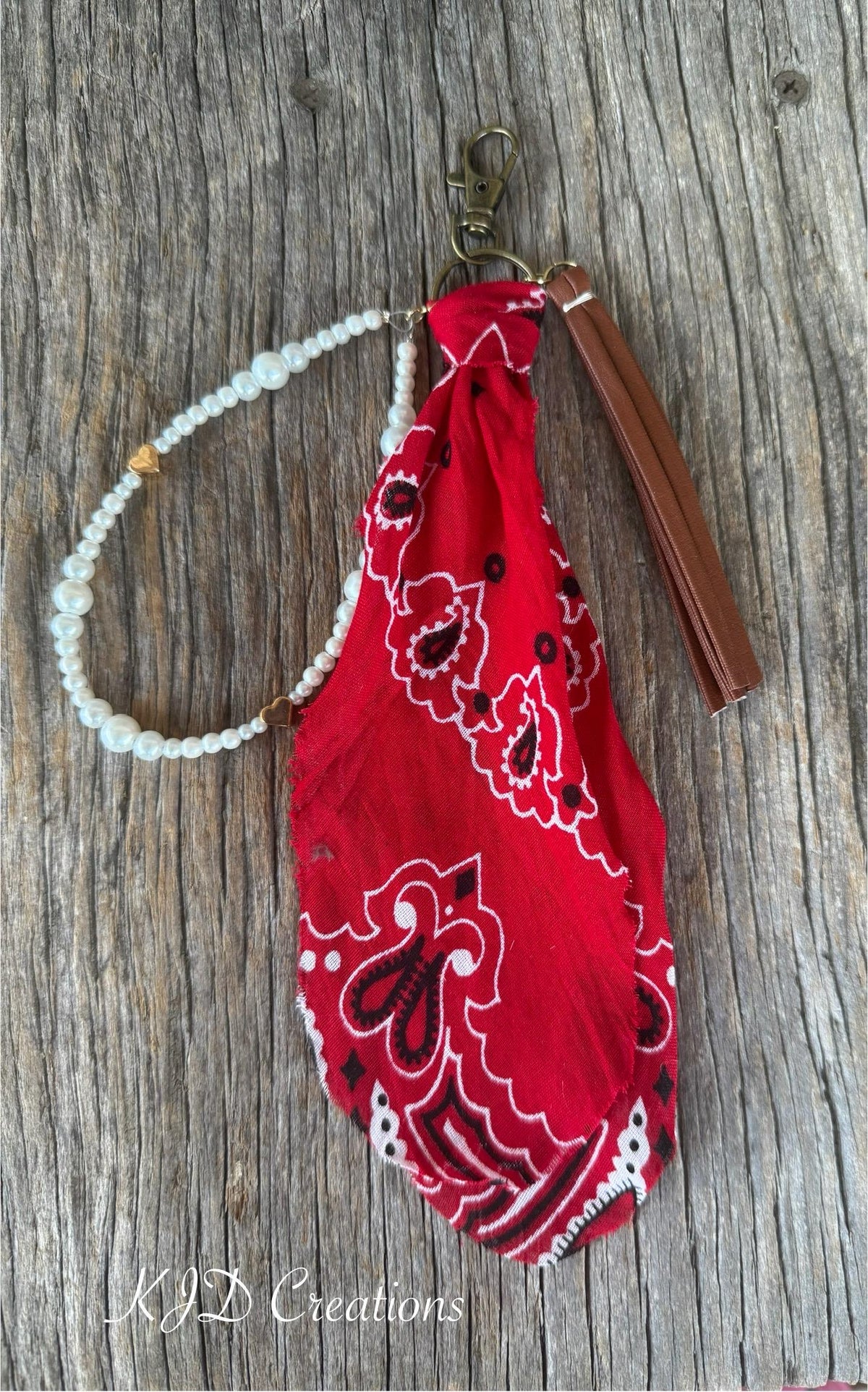 Flat lay of red paisley bandana bag charm on weathered wood showing gold lobster clasp and ring, red bandana, white faux pearl bead strand with gold heart-shaped bead accent, and brown leather fringe tassel, Western boho purse keychain charm