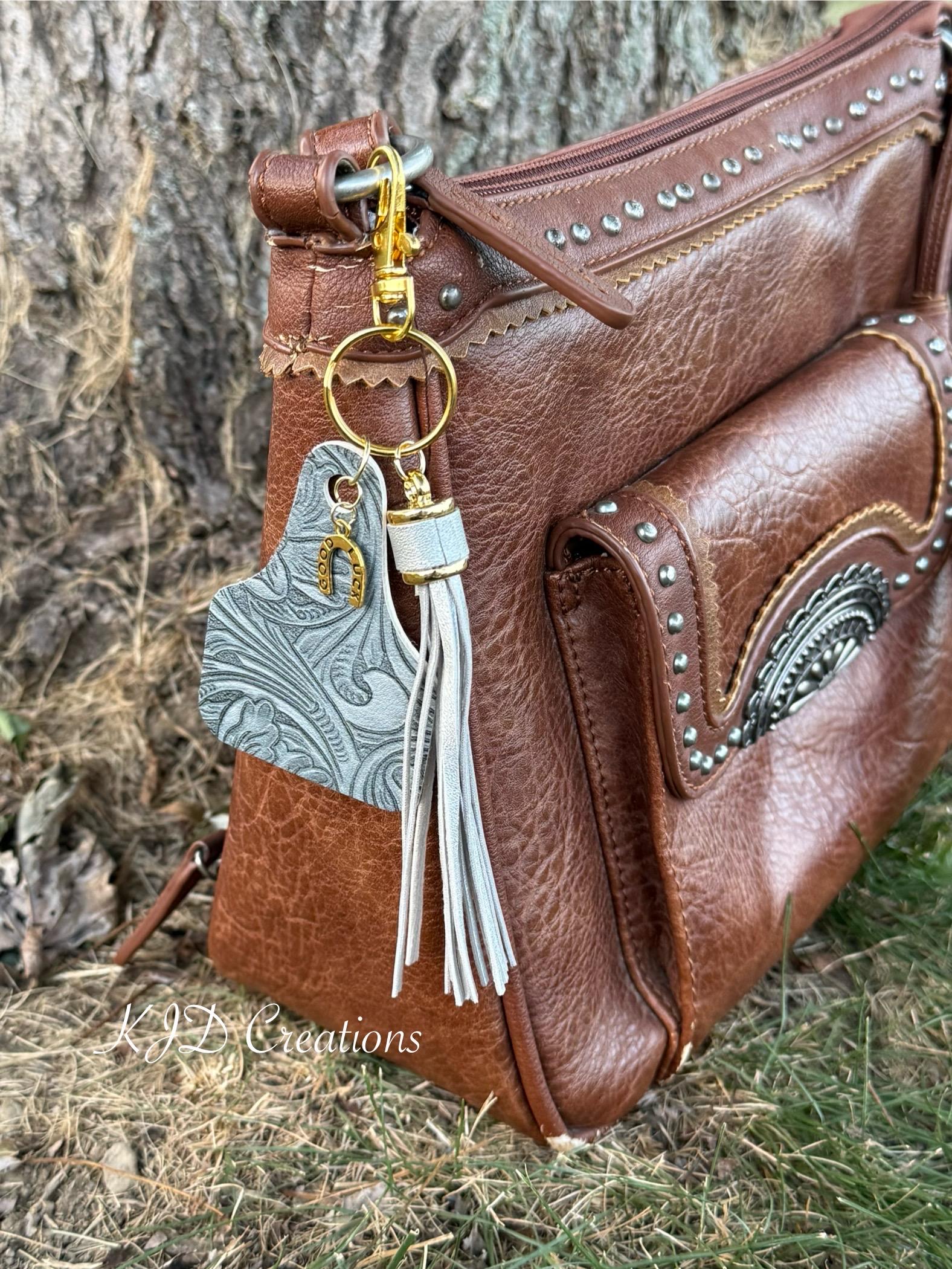 Gray tooled leather cow tag keychain bag charm with leather fringe tassel and gold "Good Luck" horseshoe charm clipped to brown studded leather handbag on weathered wood, Western boho purse accessory