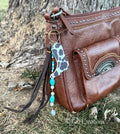 Side view of brown studded Western leather crossbody bag against tree with boho Western bag charm featuring black and white cow print faux leather cowboy boot, turquoise heishi and oval bead strand with white round beads, and dark brown leather fringe tassel, KJD Creations