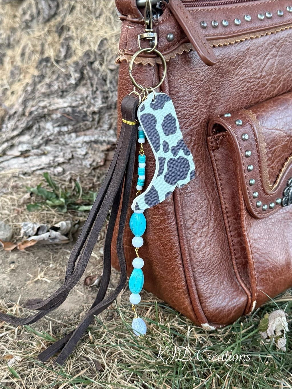 Front view of brown studded Western leather crossbody bag against tree with boho Western bag charm featuring black and white cow print faux leather cowboy boot charm, turquoise bead strand, and dark brown leather fringe tassel clipped to zipper hardware