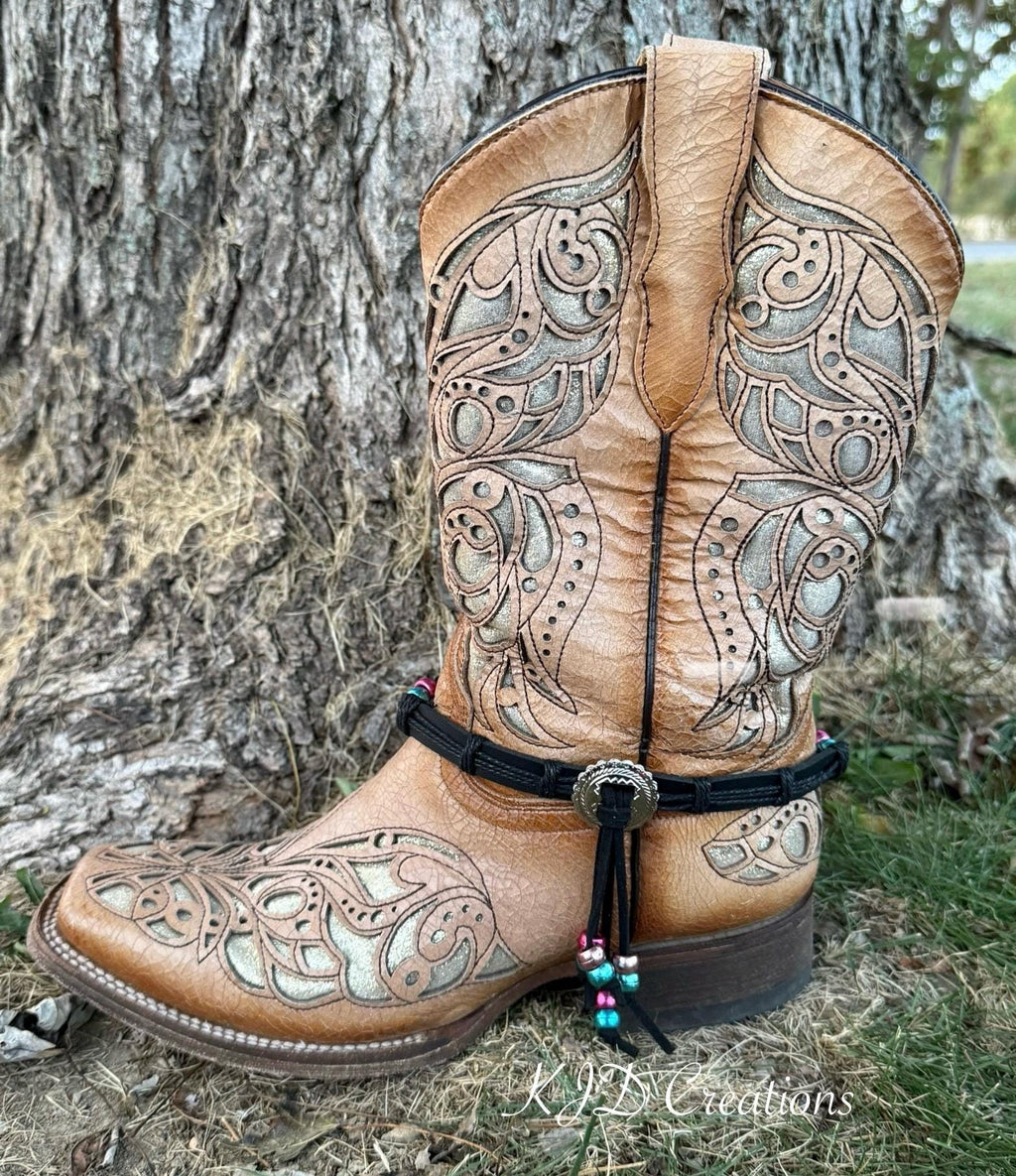 A tan leather western boot with intricate silver-toned tooled embroidery, a black ankle strap with a silver concho, and a beaded tassel, photographed outdoors against a tree trunk.