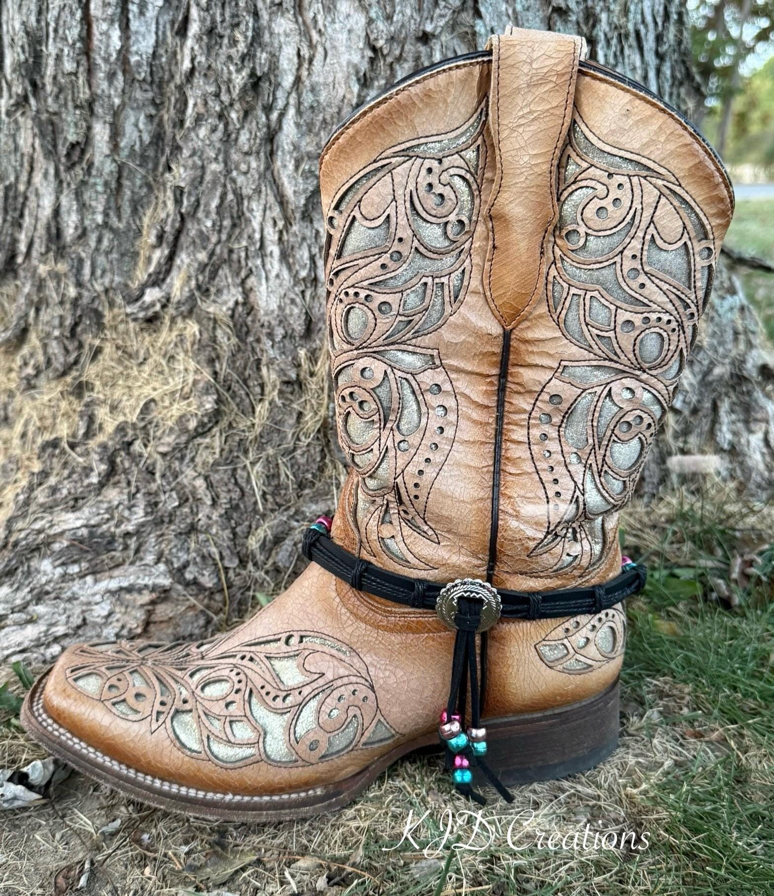 A tan leather western boot with intricate silver-toned tooled embroidery, a black ankle strap with a silver concho, and a beaded tassel, photographed outdoors against a tree trunk.