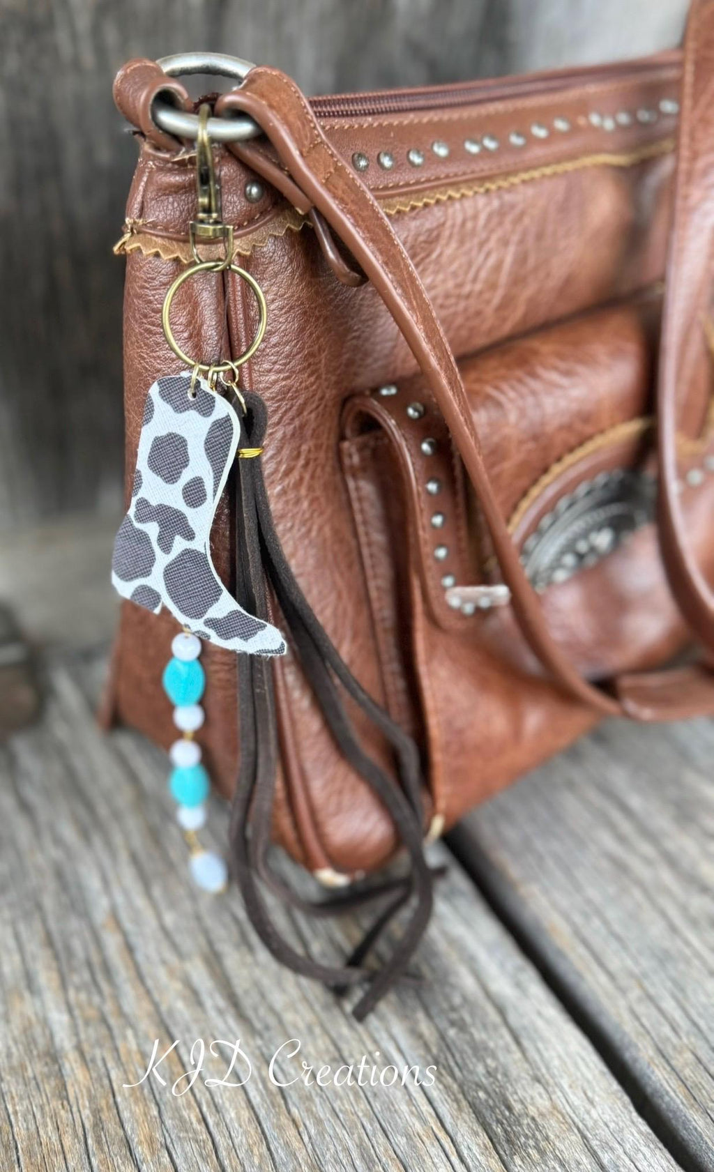Side view of brown studded Western leather crossbody bag on weathered wood with boho Western bag charm featuring black and white cow print faux leather cowboy boot, turquoise bead strand, and dark brown leather fringe tassel, Western boho purse charm