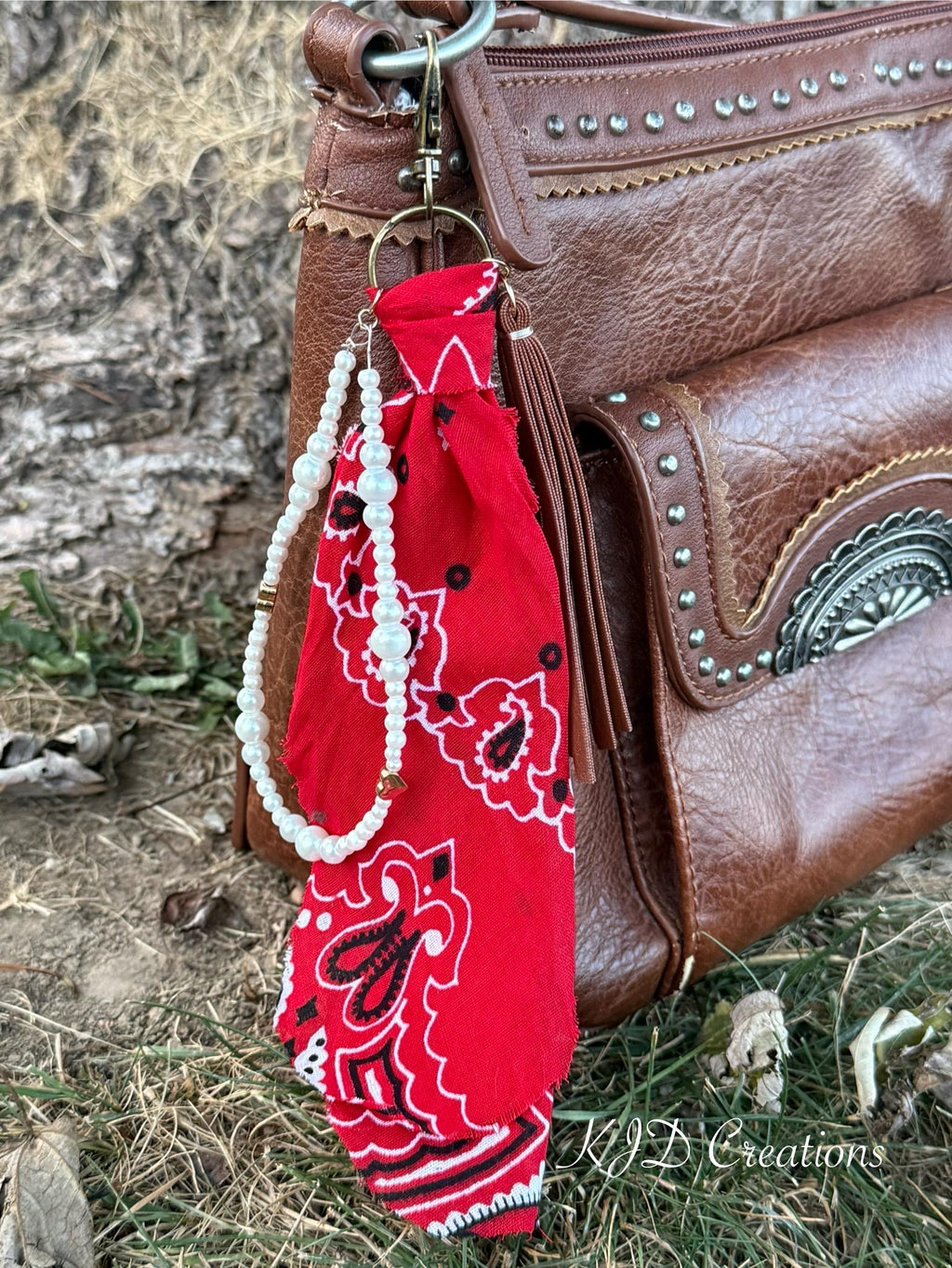 Close-up of red paisley bandana bag charm on brown studded Western leather crossbody bag showing gold ring clip, red bandana, white faux pearl bead strand with gold heart-shaped bead accent, and brown leather fringe tassel, Western boho purse keychain