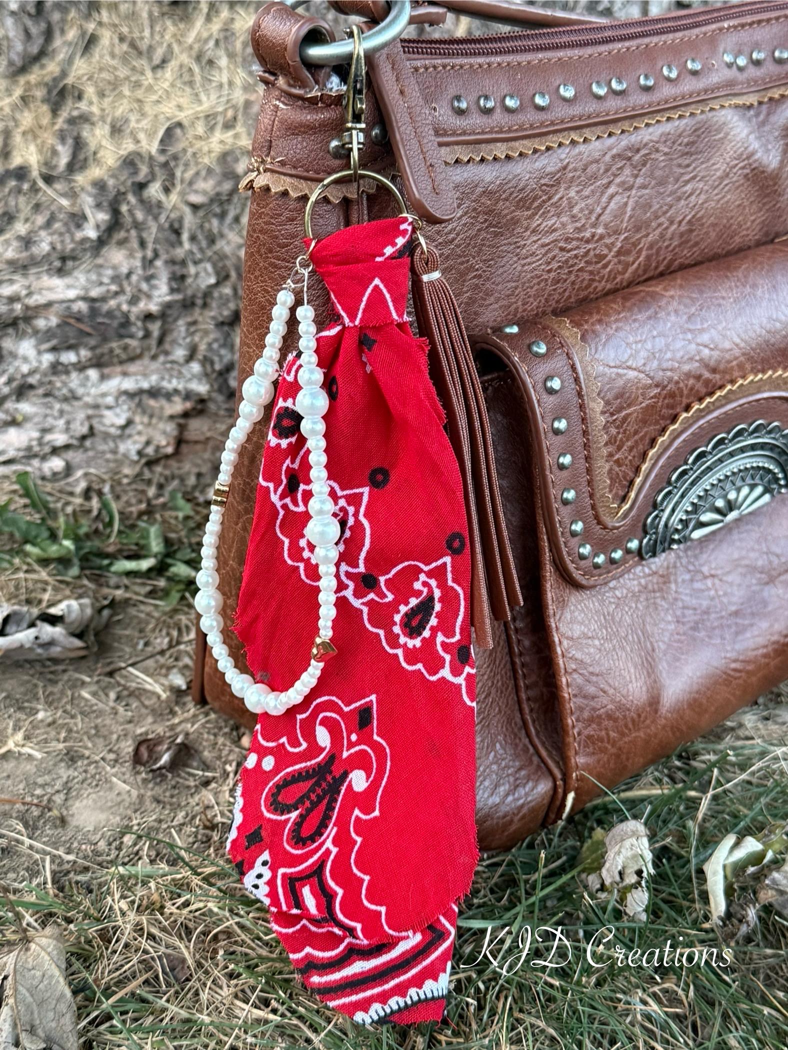 Close-up of red paisley bandana bag charm on brown studded Western leather crossbody bag showing gold ring clip, red bandana, white faux pearl bead strand with gold heart-shaped bead accent, and brown leather fringe tassel, Western boho purse keychain