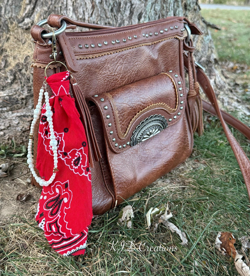 Side view of brown studded Western leather crossbody bag against tree with red paisley bandana bag charm featuring white faux pearl bead strand with gold heart-shaped bead accent and brown leather fringe tassel clipped to zipper hardware, KJD Creations Western boho purse charm