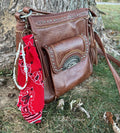 Side view of brown studded Western leather crossbody bag against tree with red paisley bandana bag charm featuring white faux pearl bead strand with gold heart-shaped bead accent and brown leather fringe tassel clipped to zipper hardware, KJD Creations Western boho purse charm