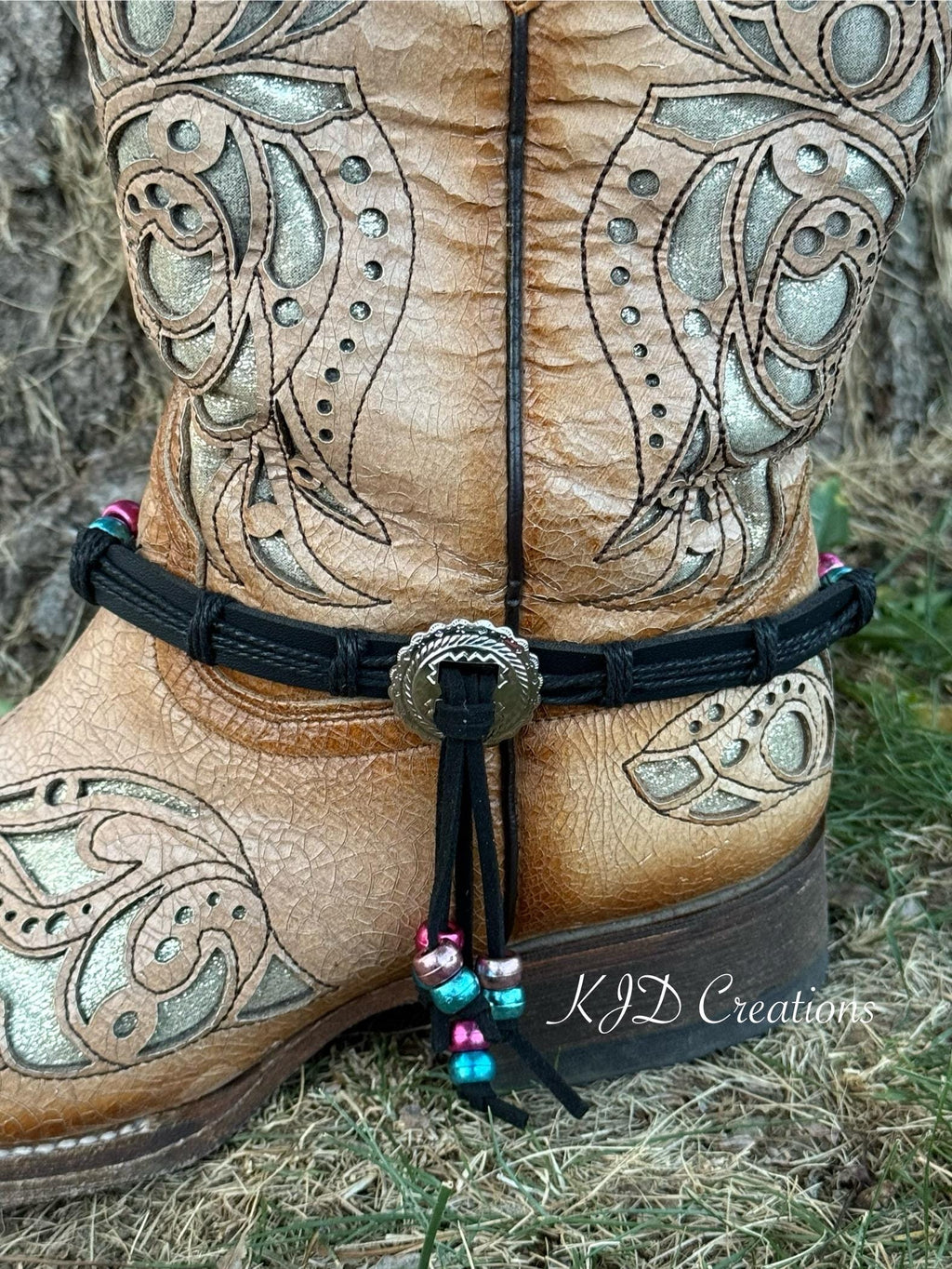 A close-up of a tan leather western boot with ornate silver-toned tooling, featuring a black ankle strap adorned with a silver concho and colorful beads, set outdoors on dry grass.