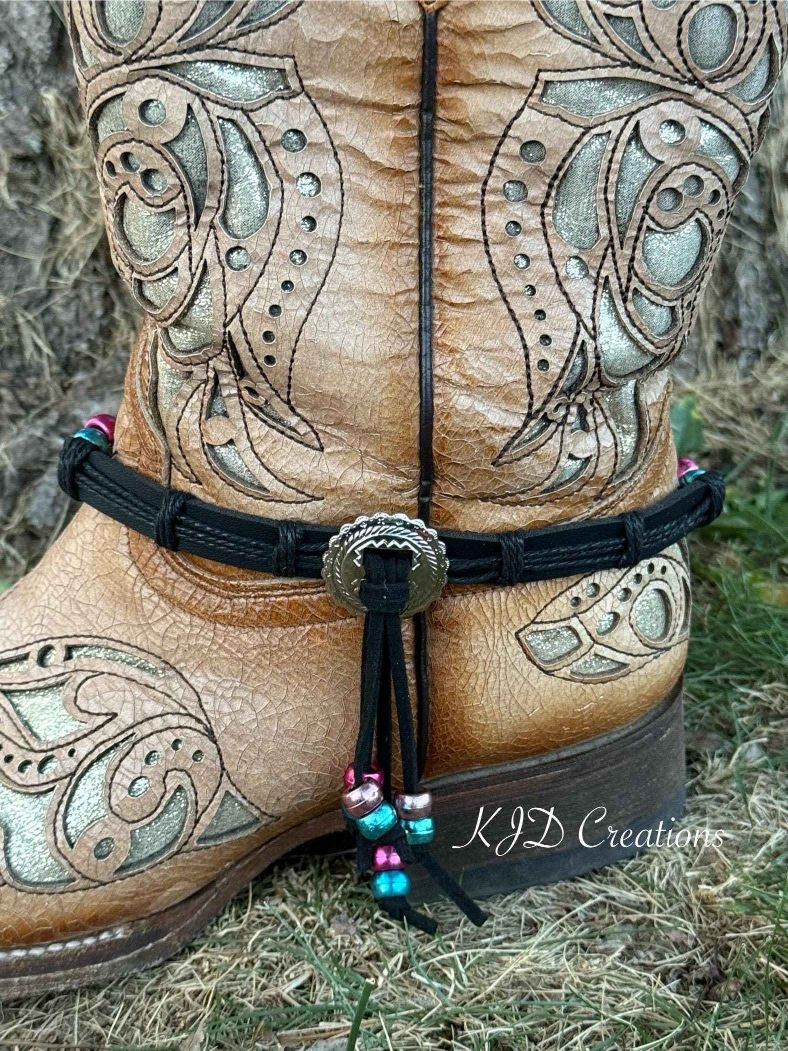 A close-up of a tan leather western boot with ornate silver-toned tooling, featuring a black ankle strap adorned with a silver concho and colorful beads, set outdoors on dry grass.