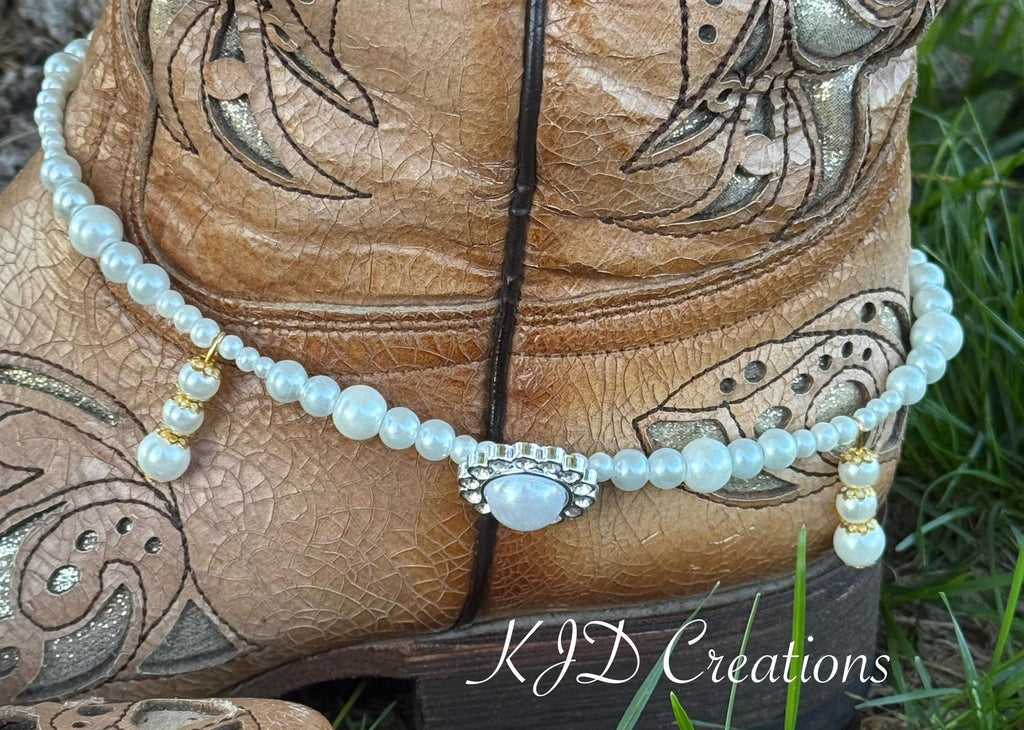 Macro close-up of pearl beaded boot bracelet with rhinestone and pearl oval concho centerpiece draped over tan tooled leather cowboy boot, bridal Western wedding accessory