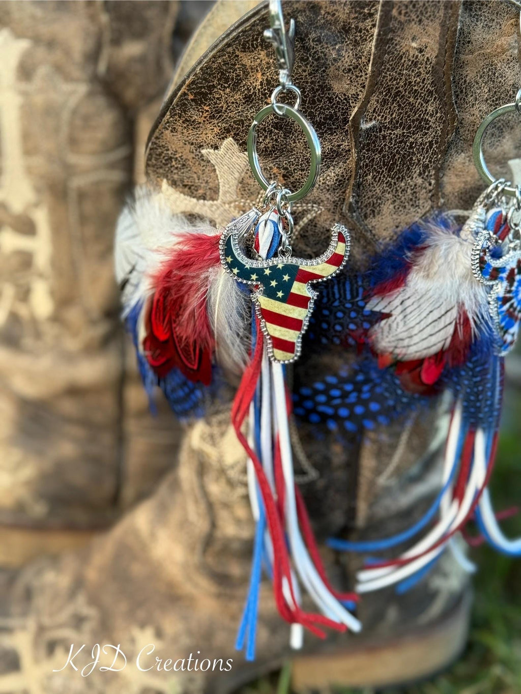 Close-up of Stars & Stripes Western boho purse charm hanging from brown distressed cowboy boot showing rhinestone-bordered American flag longhorn steer charm, red white and blue marabou and guinea fowl spotted feather bundle, and red white and blue suede leather fringe tassel strands on silver key ring