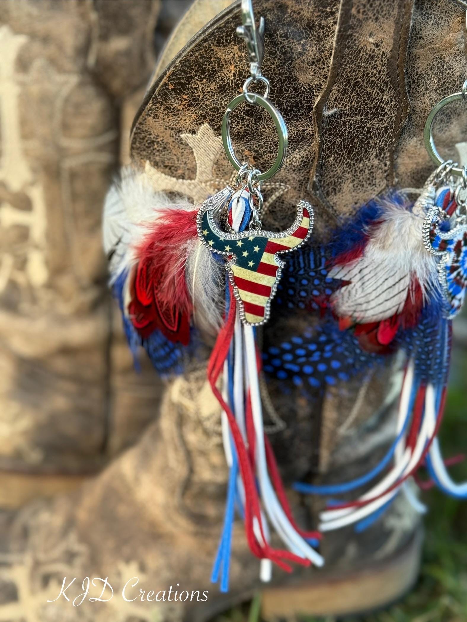 Close-up of Stars & Stripes Western boho purse charm hanging from brown distressed cowboy boot showing rhinestone-bordered American flag longhorn steer charm, red white and blue marabou and guinea fowl spotted feather bundle, and red white and blue suede leather fringe tassel strands on silver key ring