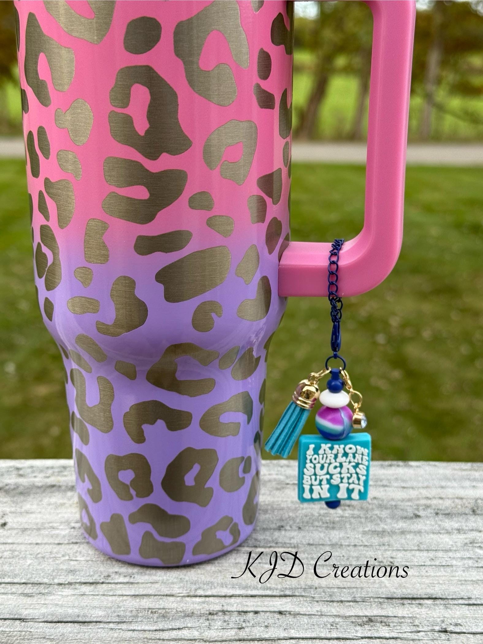 Stay In Your Lane funny tumbler cup handle charm clipped to a pink and purple leopard print insulated tumbler — navy blue chain with teal tassel, tie-dye bead, and teal square charm reading "I Know Your Lane Sucks But Stay In It" by KJD Creations