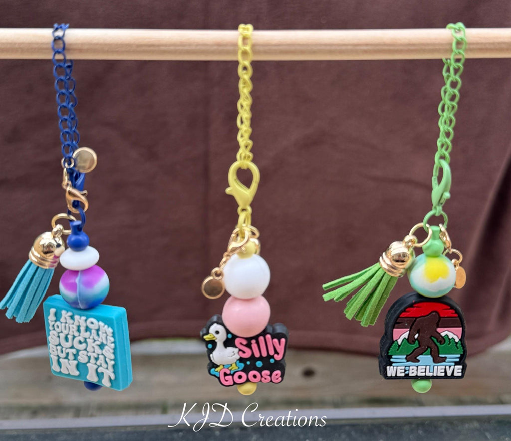 Three funny tumbler cup handle charms hanging on a display rack — Stay In Your Lane teal square charm with blue chain, Silly Goose penguin charm with yellow chain, and We Believe Bigfoot charm with green chain by KJD Creations