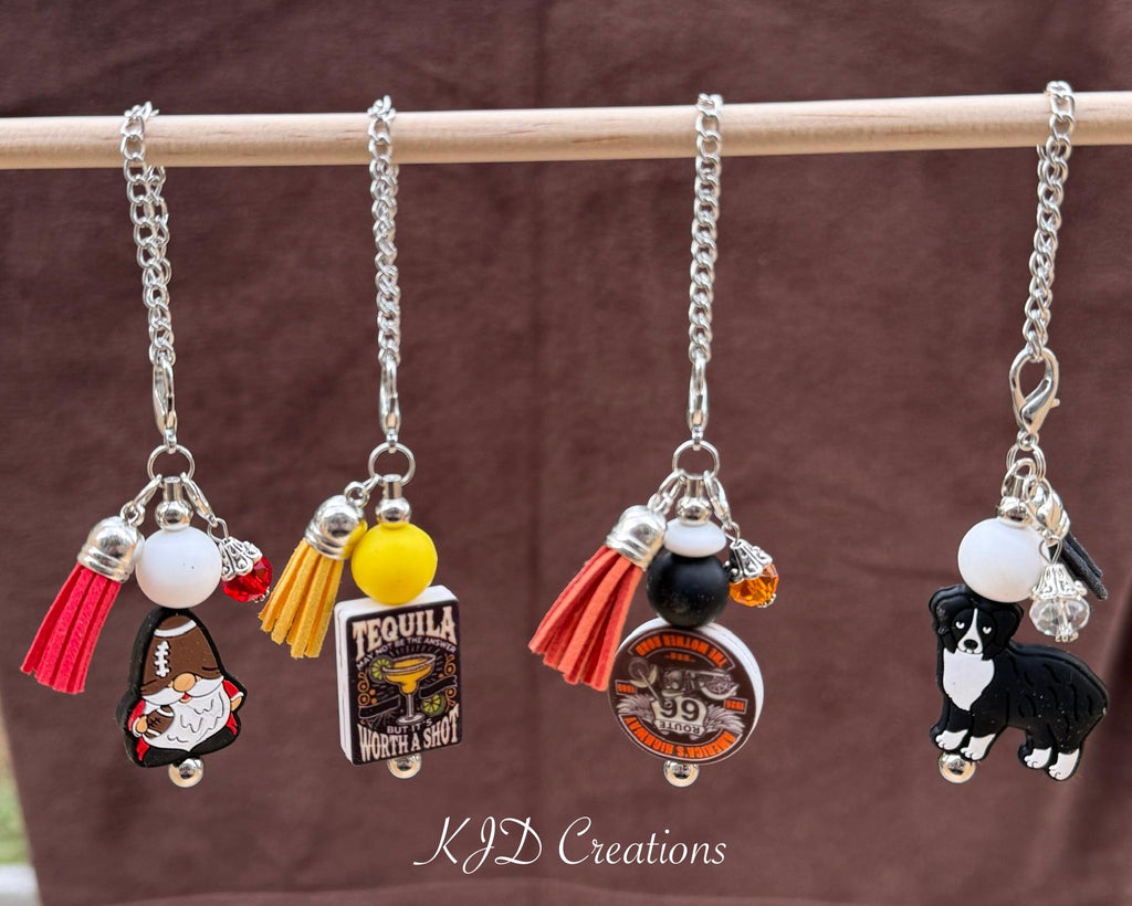 Four silicone beaded tumbler dangle charms hanging on a wood display rack — Football Gnome with red tassel, Tequila Worth a Shot with yellow tassel, Route 66 with orange tassel, and Aussie dog charm with white bead, all on silver chains by KJD Creations