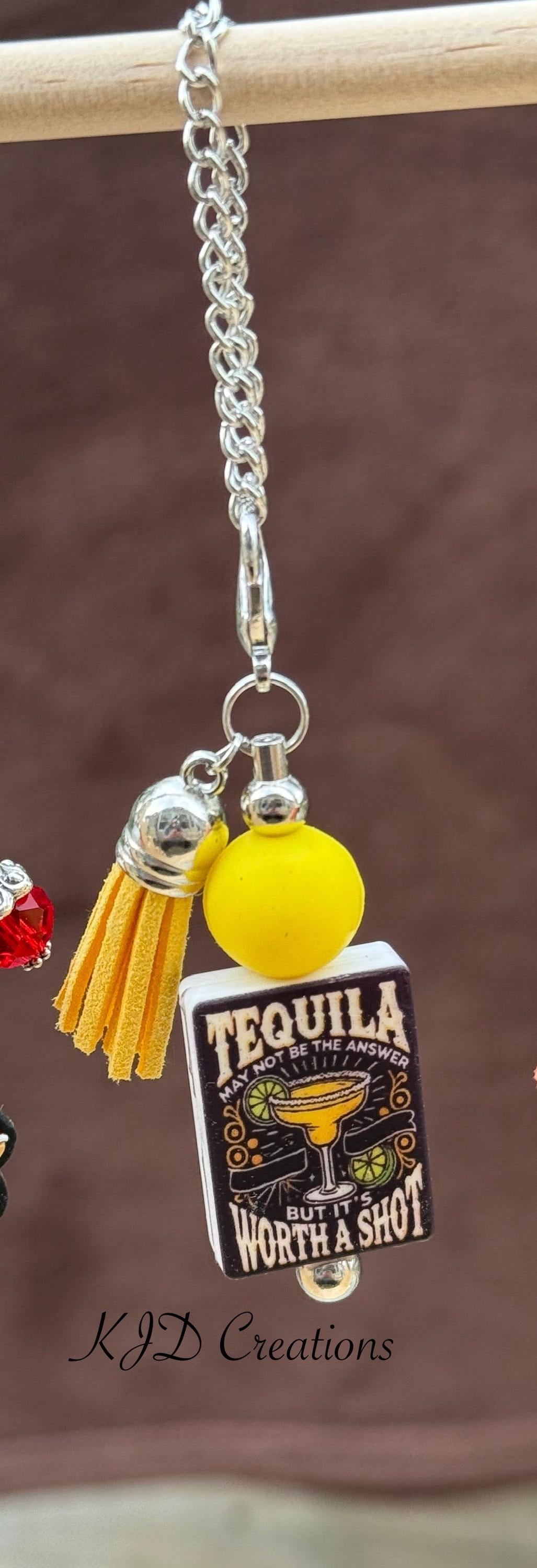 Tequila Worth a Shot tumbler dangle charm close-up on display rack — silver chain with yellow silicone bead, gold leather tassel, and black square "Tequila May Not Be the Answer But It's Worth a Shot" charm by KJD Creations