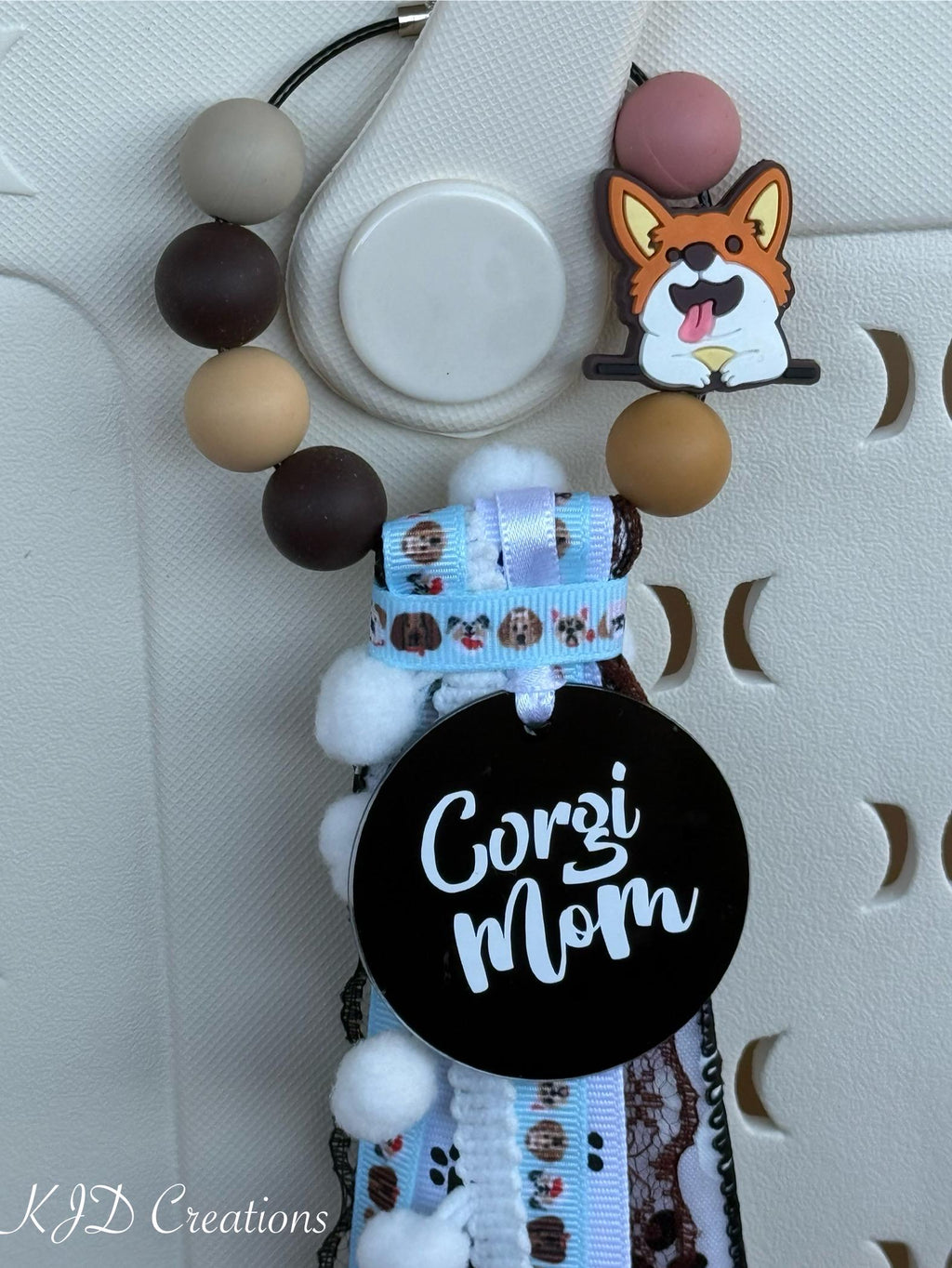 Alternate close-up of Corgi Mom silicone beaded bag tassel on white Hurley tote bag showing earth-tone round silicone bead loop with Corgi silicone dog charm, large black round Corgi Mom acrylic pendant with white script, white pom-pom trim bead strand, and cascading light blue dog-print grosgrain, black and white pawprint grosgrain, and white pom-pom trim ribbon tassel, KJD Creations
