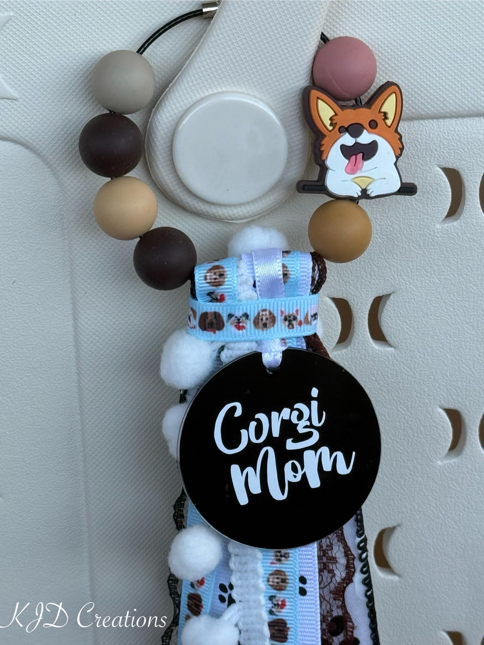 Alternate close-up of Corgi Mom silicone beaded bag tassel on white Hurley tote bag showing earth-tone round silicone bead loop with Corgi silicone dog charm, large black round Corgi Mom acrylic pendant with white script, white pom-pom trim bead strand, and cascading light blue dog-print grosgrain, black and white pawprint grosgrain, and white pom-pom trim ribbon tassel, KJD Creations