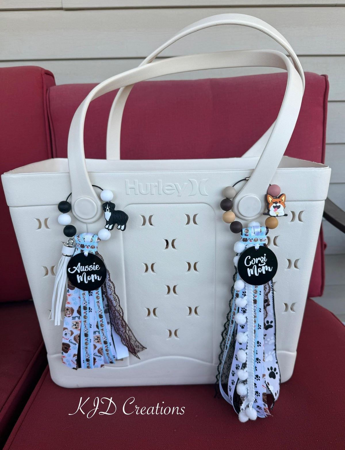 Both Aussie Mom and Corgi Mom silicone beaded bag tassels clipped to white Hurley tote bag handles on red chair — Aussie Mom tassel on left with black and white silicone bead loop, Australian Shepherd charm, black Aussie Mom acrylic pendant, and dog-print ribbon tassel; Corgi Mom tassel on right with earth-tone silicone bead loop, Corgi charm, black Corgi Mom acrylic pendant, and pawprint ribbon tassel with white pom-pom trim, KJD Creations