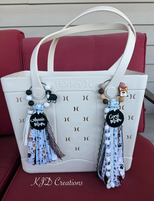 Both Aussie Mom and Corgi Mom silicone beaded bag tassels clipped to white Hurley tote bag handles on red chair — Aussie Mom tassel on left with black and white silicone bead loop, Australian Shepherd charm, black Aussie Mom acrylic pendant, and dog-print ribbon tassel; Corgi Mom tassel on right with earth-tone silicone bead loop, Corgi charm, black Corgi Mom acrylic pendant, and pawprint ribbon tassel with white pom-pom trim, KJD Creations