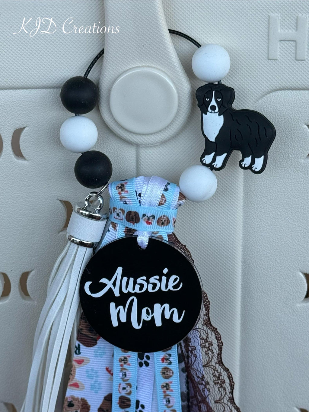 Alternate close-up of Aussie Mom silicone beaded bag tassel on white Hurley tote bag showing black and white round silicone bead loop with Australian Shepherd silicone dog charm, large black round Aussie Mom acrylic pendant with white script, and cascading light blue dog-print grosgrain, white leather fringe, and black lace ribbon tassel, KJD Creations