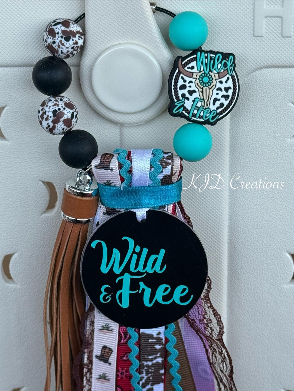 Close-up of Wild & Free bag tassel with black charm disc, turquoise silicone beads, cow print charm, and colorful Western ribbon fringe on a Bogg-style bag