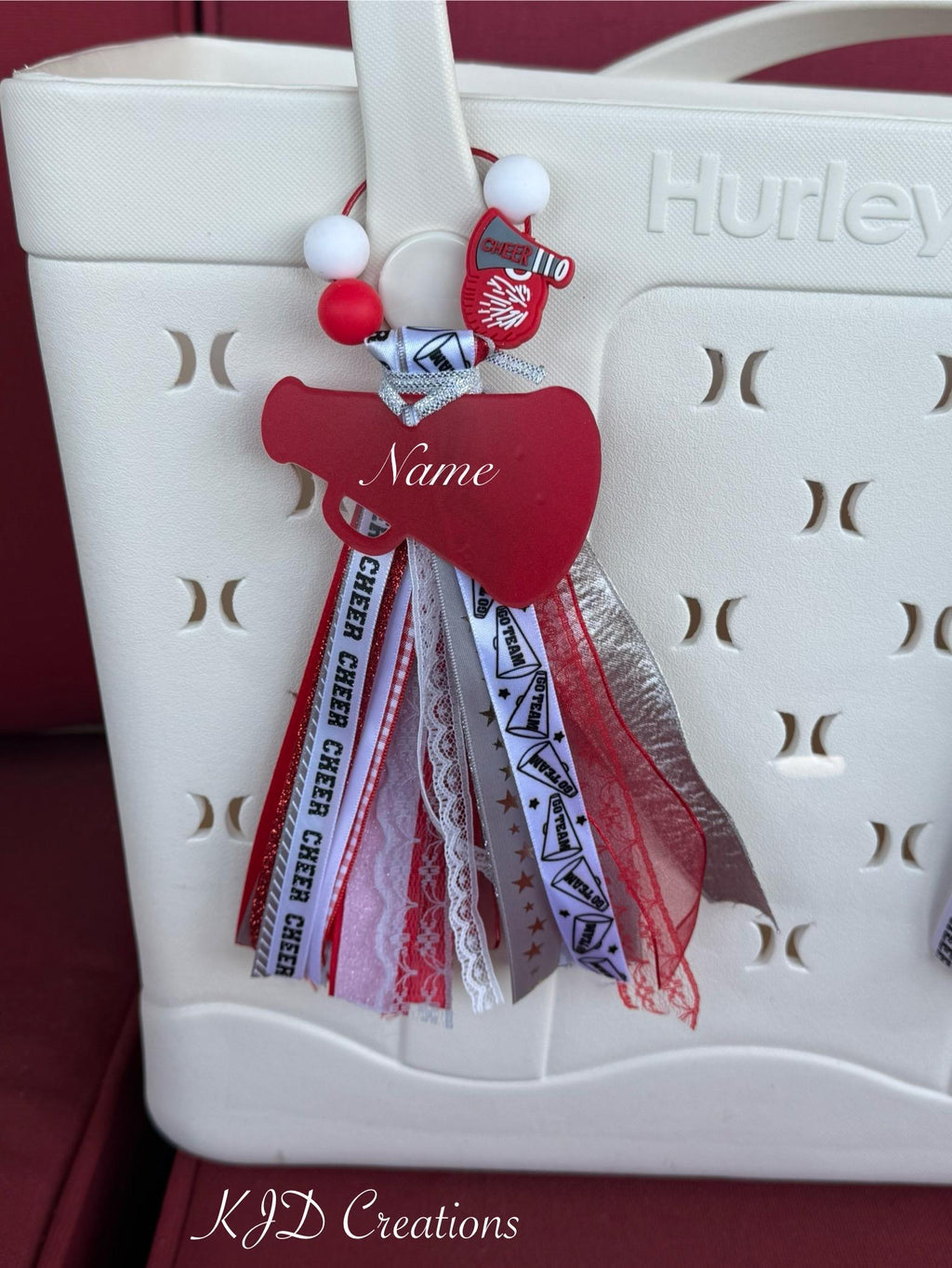 Red personalized cheer silicone beaded bag tassel clipped to white Hurley tote bag handle against red background, featuring wire loop with red and white silicone beads and red Cheer 110 megaphone silicone charm, silver glitter ribbon bow, large red acrylic megaphone-heart pendant with personalized name in white script, and cascading red and white Cheer print grosgrain white lace red lace silver glitter Go Team print and red grosgrain ribbon tassel, KJD Creations