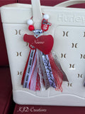 Red personalized cheer silicone beaded bag tassel clipped to white Hurley tote bag handle against red background, featuring wire loop with red and white silicone beads and red Cheer 110 megaphone silicone charm, silver glitter ribbon bow, large red acrylic megaphone-heart pendant with personalized name in white script, and cascading red and white Cheer print grosgrain white lace red lace silver glitter Go Team print and red grosgrain ribbon tassel, KJD Creations