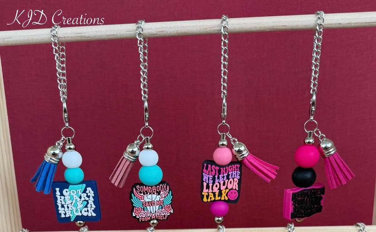 Four country music tumbler cup handle charms displayed on a rack — Truck, Save Me, Liquor Talk, and Pink 