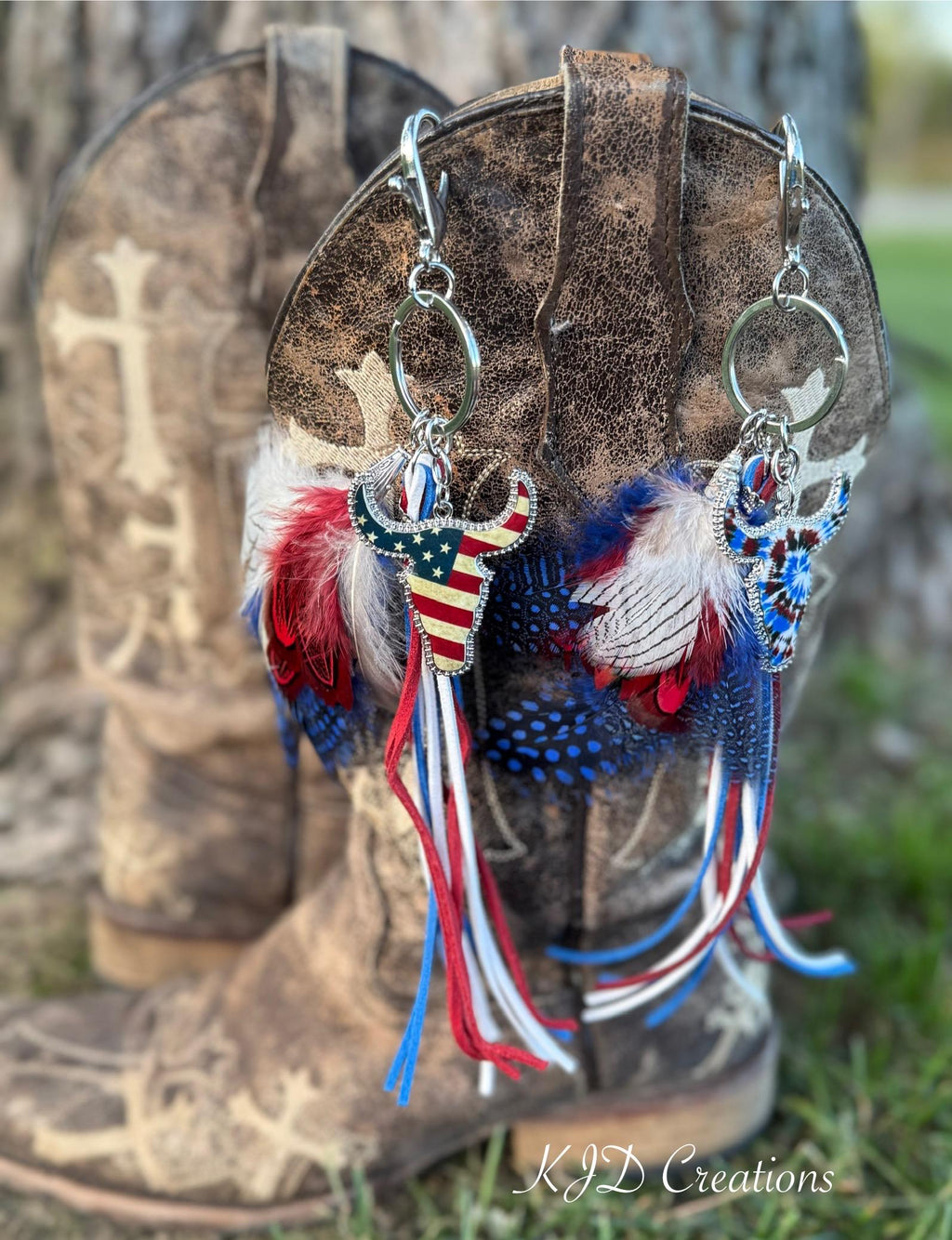 Both Western boho longhorn steer purse charm and keychain variants hanging from brown distressed cross-embossed cowboy boot outdoors — Stars & Stripes American flag longhorn steer charm on left and Tie Dye longhorn steer charm on right, both with red, white and blue feather bundles and suede fringe tassel strands, KJD Creations