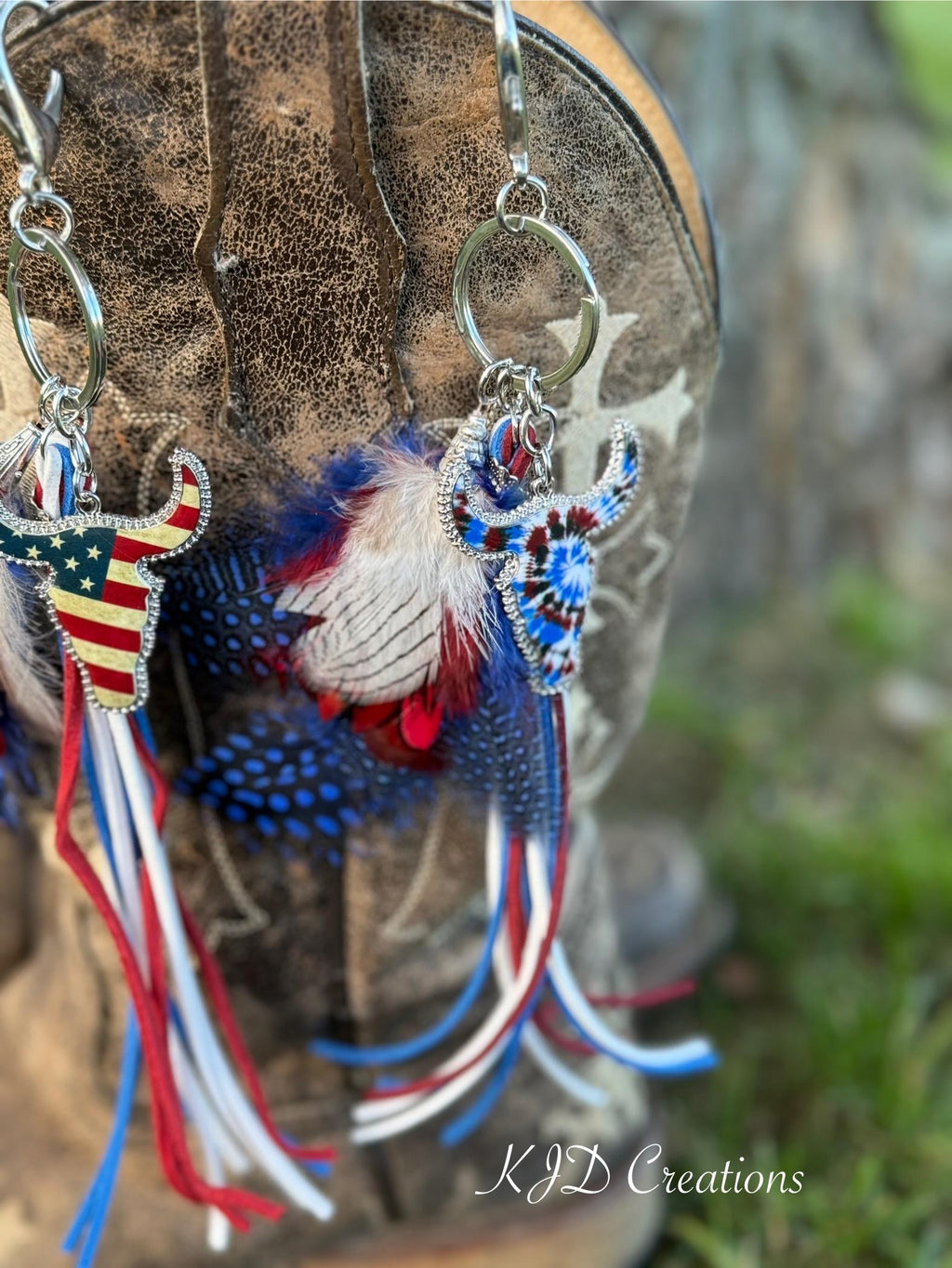 Close-up of both Western boho longhorn steer purse charm and keychain variants hanging from brown distressed cowboy boot — Stars & Stripes American flag longhorn steer charm on left and Tie Dye longhorn steer charm on right with red white and blue feather bundles and suede fringe tassel strands, KJD Creations
