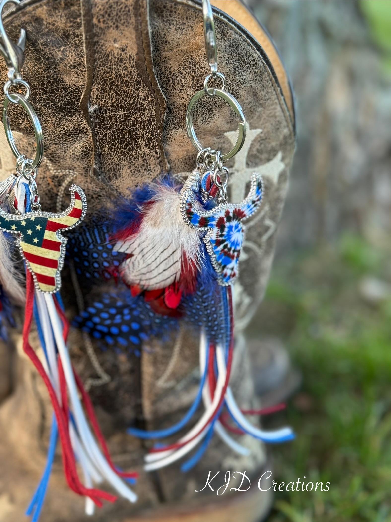 Close-up of both Western boho longhorn steer purse charm and keychain variants hanging from brown distressed cowboy boot — Stars & Stripes American flag longhorn steer charm on left and Tie Dye longhorn steer charm on right with red white and blue feather bundles and suede fringe tassel strands, KJD Creations
