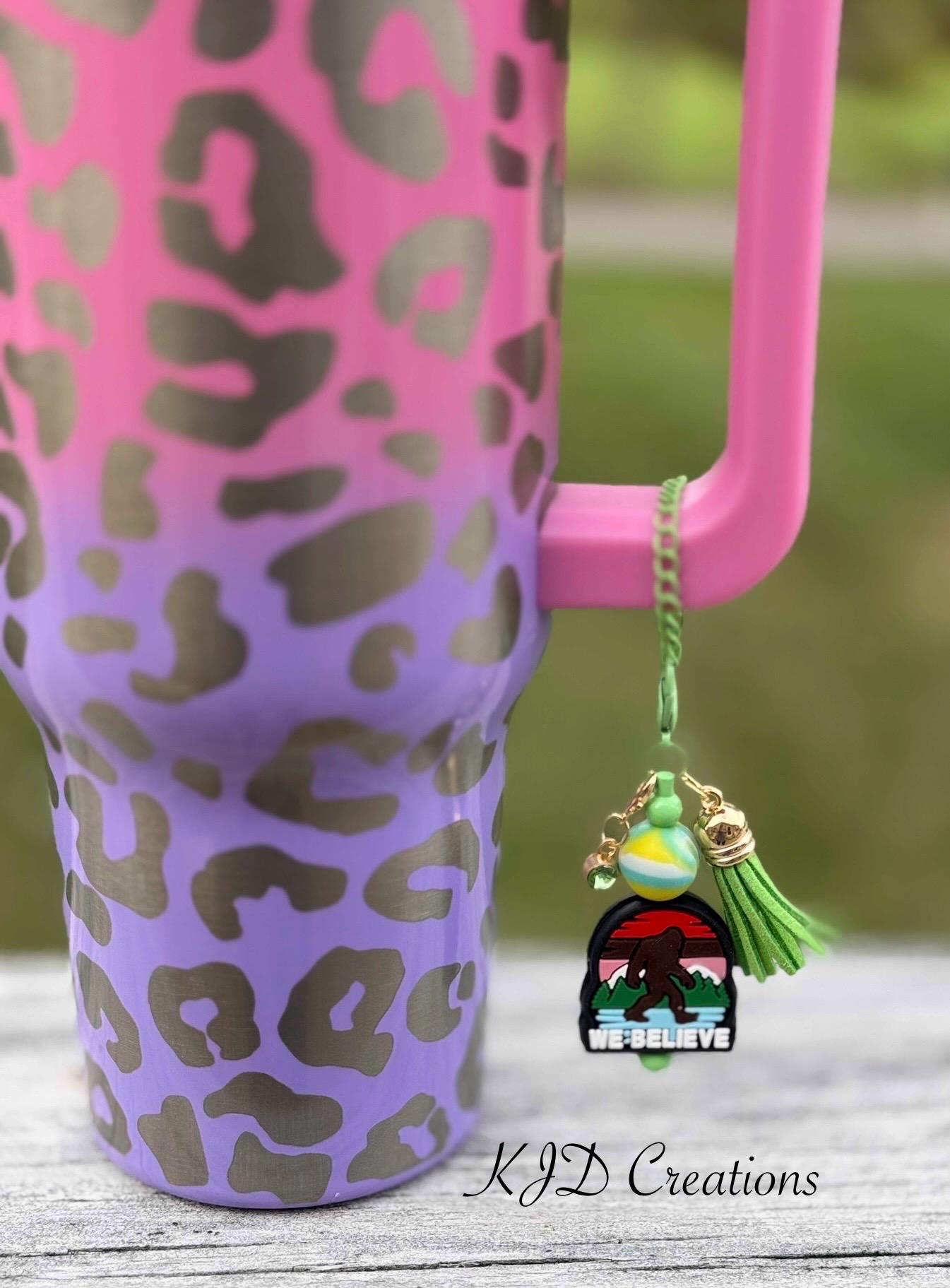 We Believe Bigfoot funny tumbler cup handle charm clipped to a pink and purple leopard print insulated tumbler — green chain with green tassel and colorful Bigfoot mountain scene charm by KJD Creations