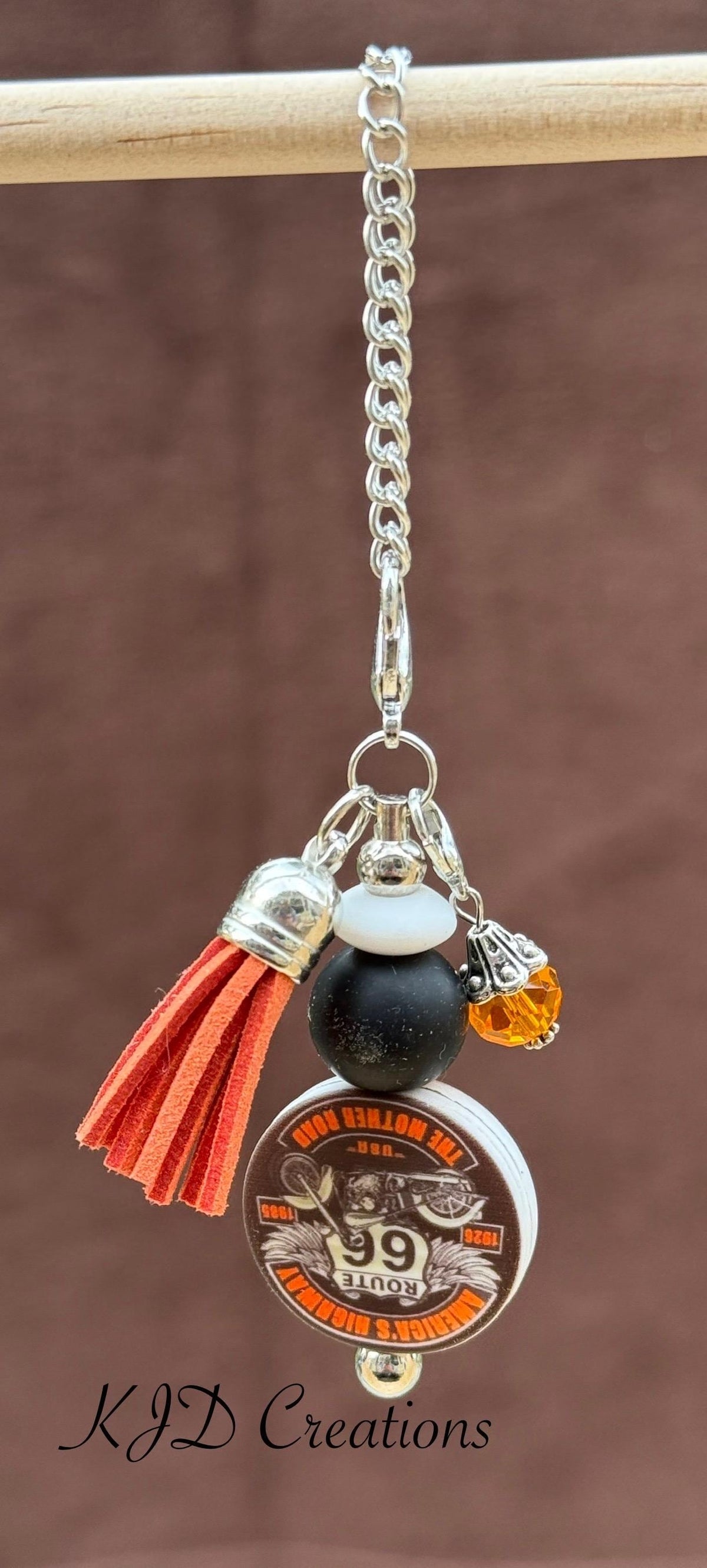 Route 66 tumbler dangle charm close-up on display rack — silver chain with white bead, black bead, amber crystal, orange leather tassel, and round Route 66 motorcycle charm by KJD Creations