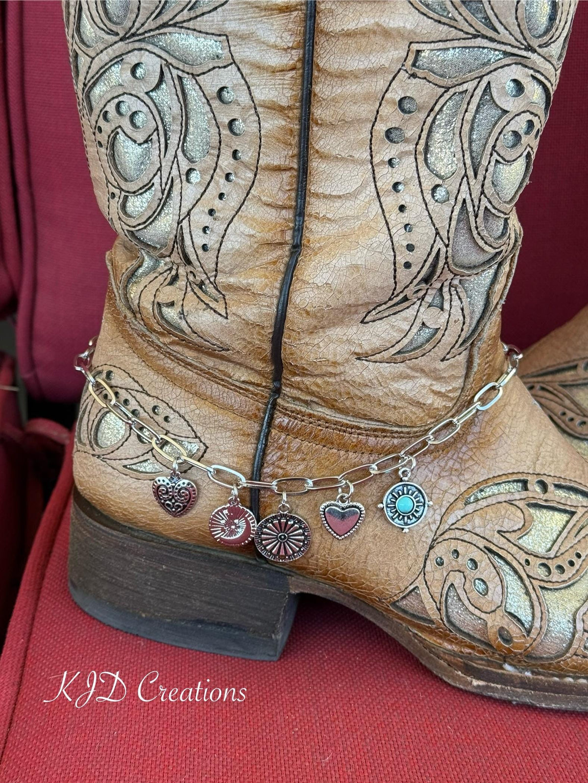 A close-up of a brown tooled leather cowboy boot with ornate western embroidery ankle with silver paperclip chain boot bracelet and five dangling Western charms including turquoise stone concho disc and silver heart and filigree heart charms