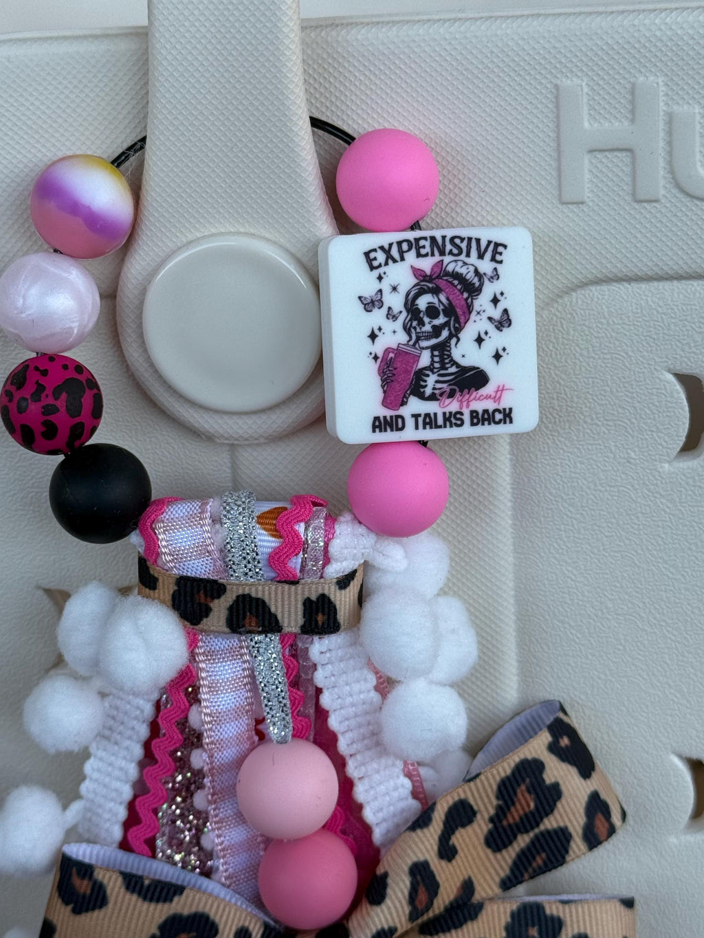 Close-up of Expensive Difficult And Talks Back silicone square bead with skeleton girl graphic, pink, black, and leopard print silicone beads, and white pom-pom trim on a bag tassel
