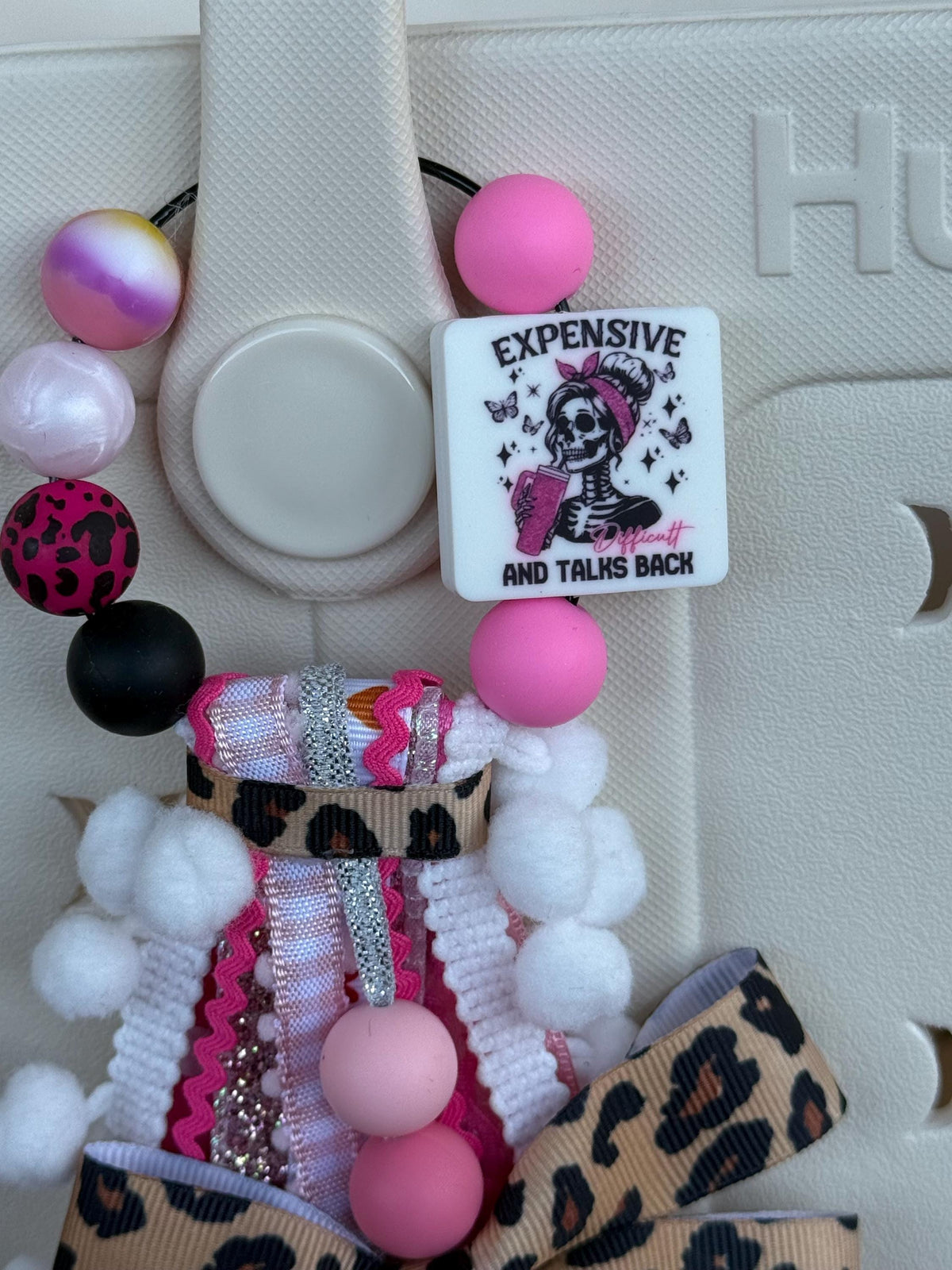 Close-up of Expensive Difficult And Talks Back silicone square bead with skeleton girl graphic, pink, black, and leopard print silicone beads, and white pom-pom trim on a bag tassel
