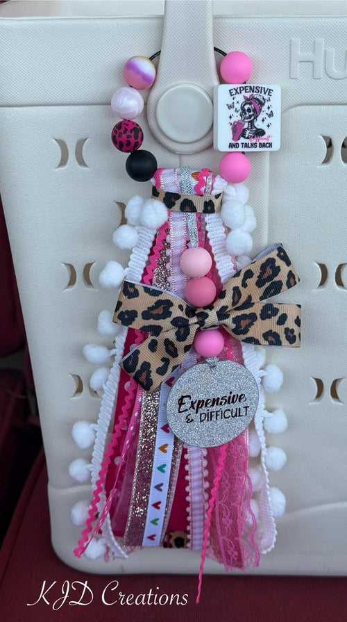 Expensive Difficult & Talks Back silicone beaded bag tassel with leopard print bow, glitter Expensive & Difficult charm disc, white pom-pom trim, and layered hot pink ribbon tassel clipped onto a white Hurley tote bag

